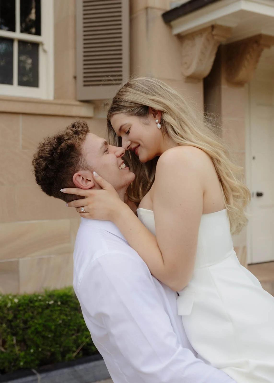 A couple in love, with the woman wearing a white dress and the man in a white shirt, holding each other closely and looking into each other's eyes outdoors in front of a building.