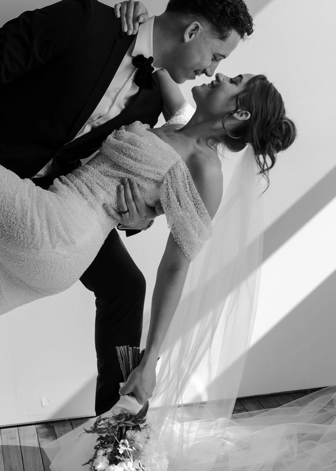 A black and white photo of a groom holding a bride, with their faces close together and smiling, in a joyful moment.