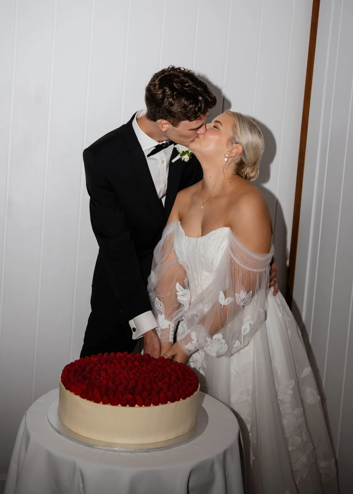 A newlywed couple kissing next to a wedding cake covered in raspberries, inside a room with white wall paneling.