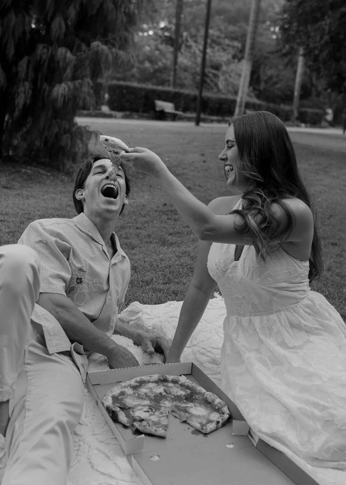 A woman playfully holding a slice of pizza near a man's mouth during a picnic outdoors, with a pizza box on a blanket and trees in the background.