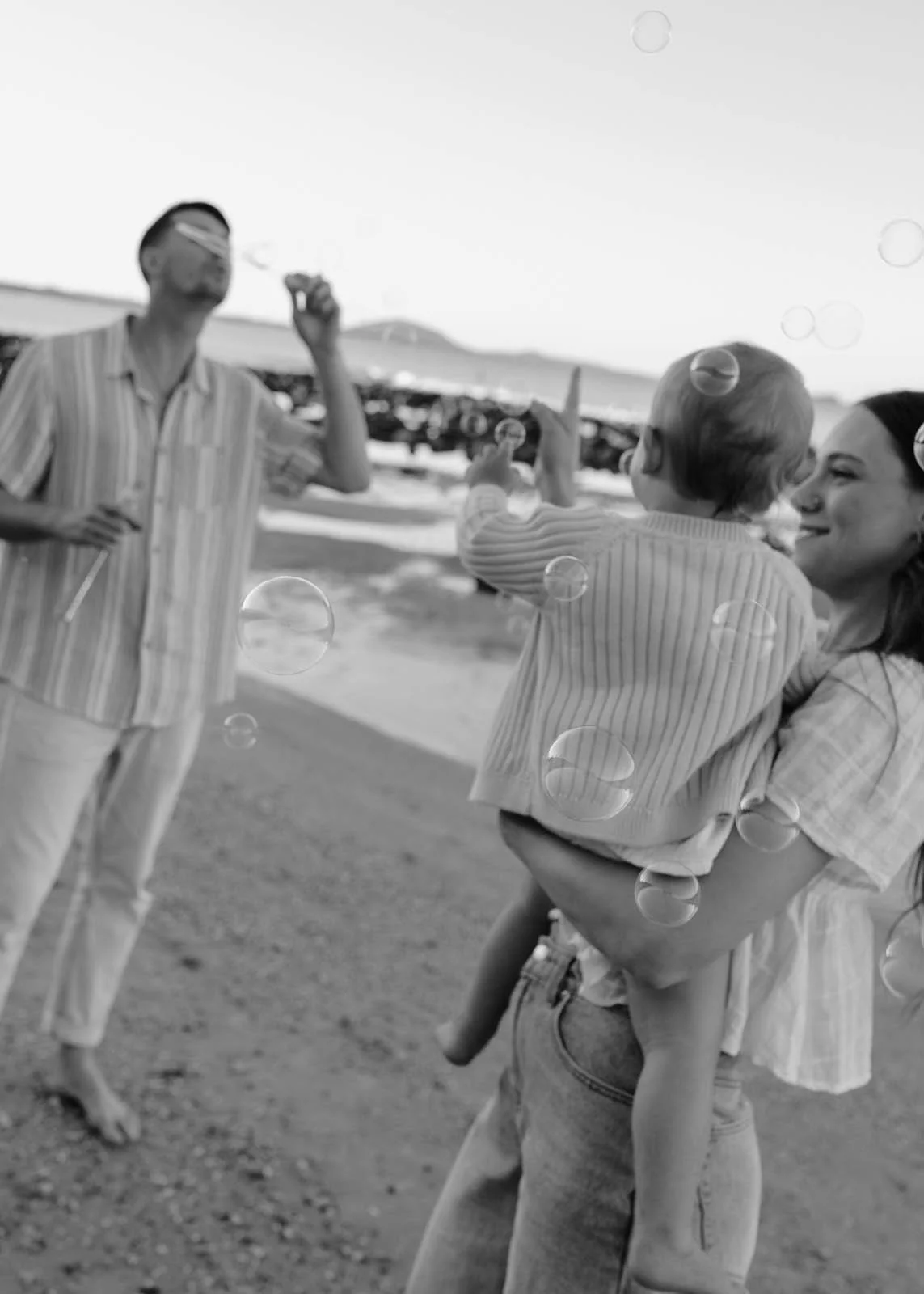 A woman holding a child on a beach, with a man in the background. The woman and child are smiling, and the child is pointing. The photo is in black and white.