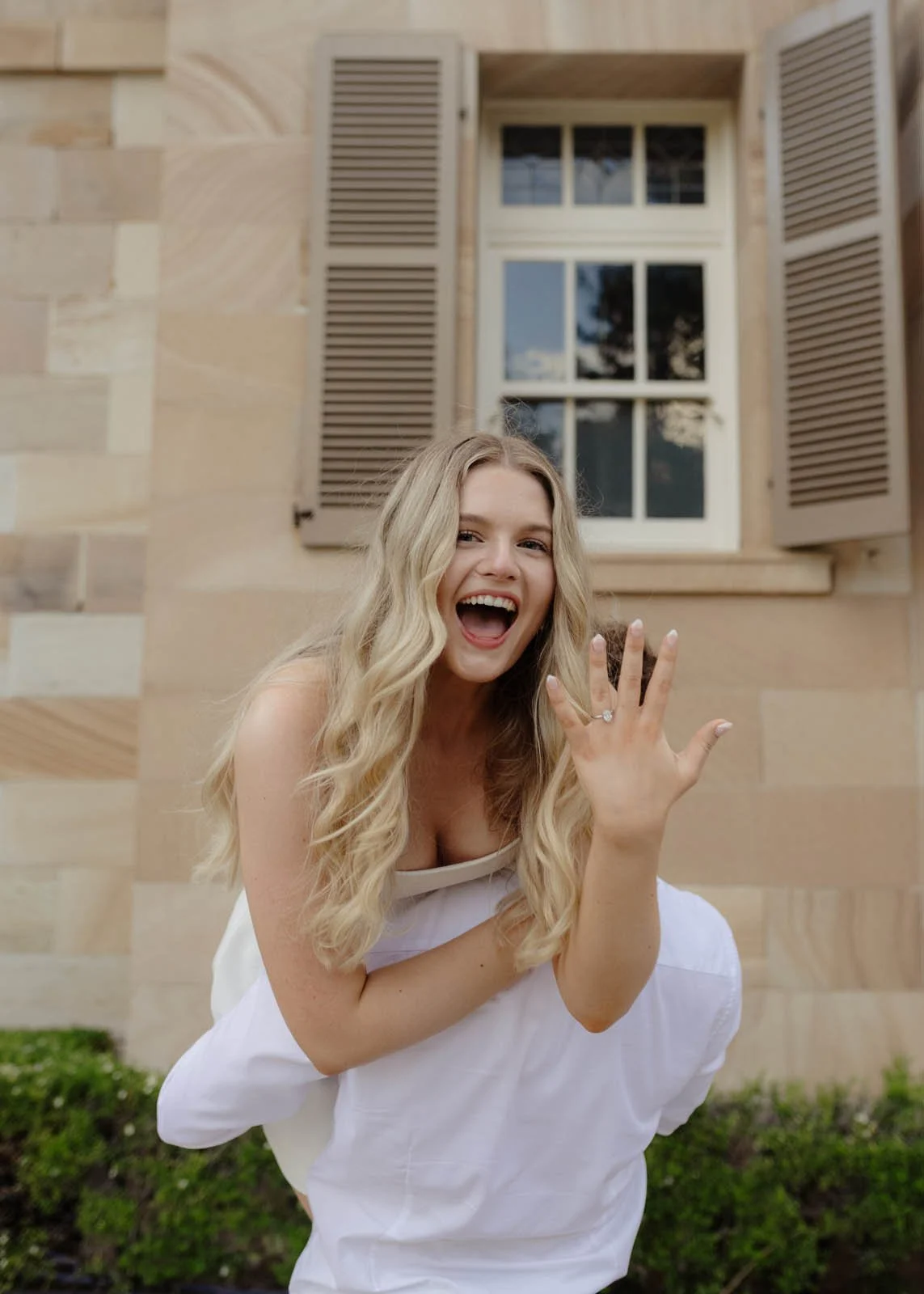 A woman with long blonde hair celebrating outdoors, showing off an engagement ring on her finger, in front of a stone building with a window and shutters.