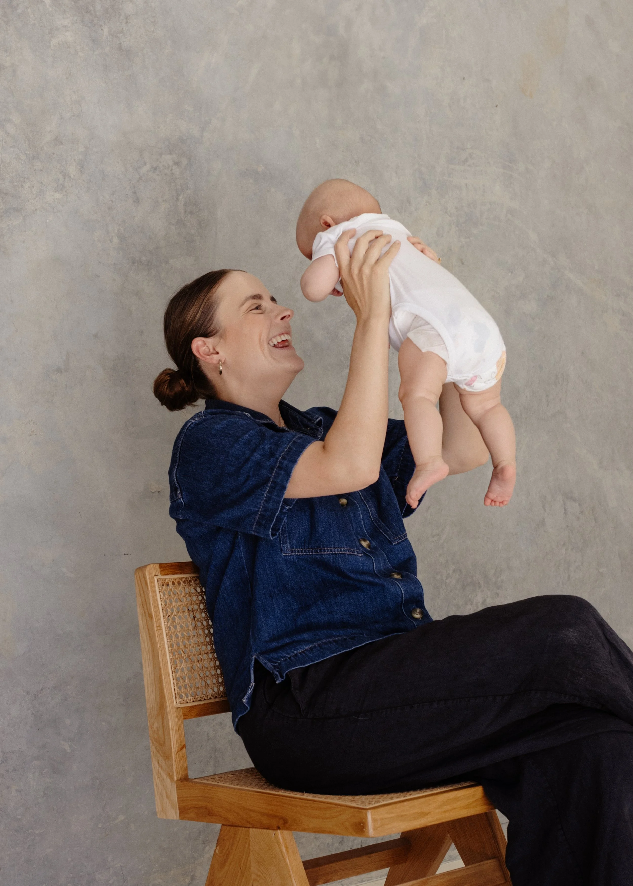 A woman with dark hair tied in a bun sitting on a wooden chair, smiling and holding a baby in the air. The woman is wearing a denim shirt and black pants, and the background is a plain textured wall.
