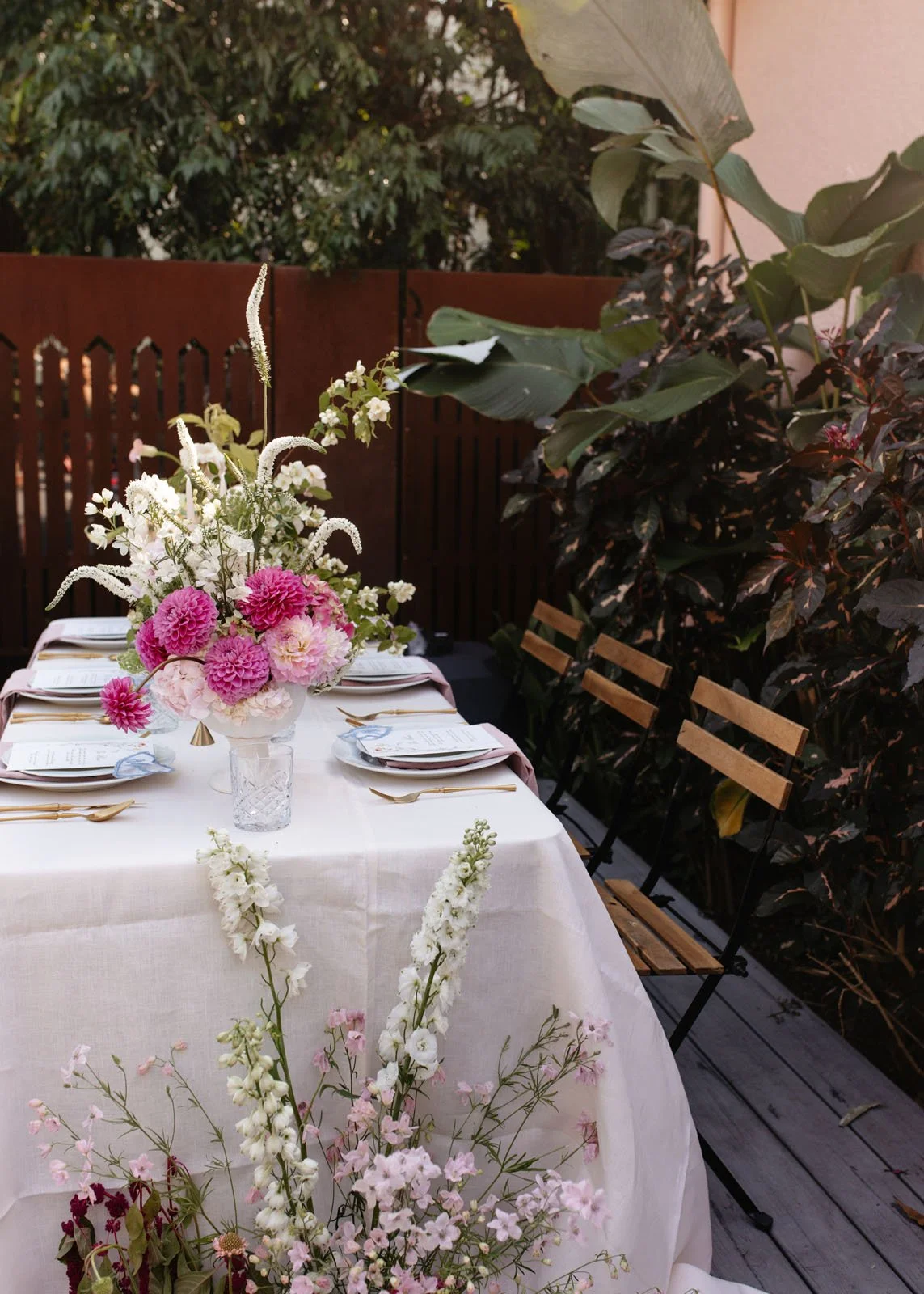 A beautifully set outdoor dining table with a white tablecloth, floral centerpiece with pink and white flowers, surrounded by wooden chairs on a wooden deck, with lush green plants and trees in the background.