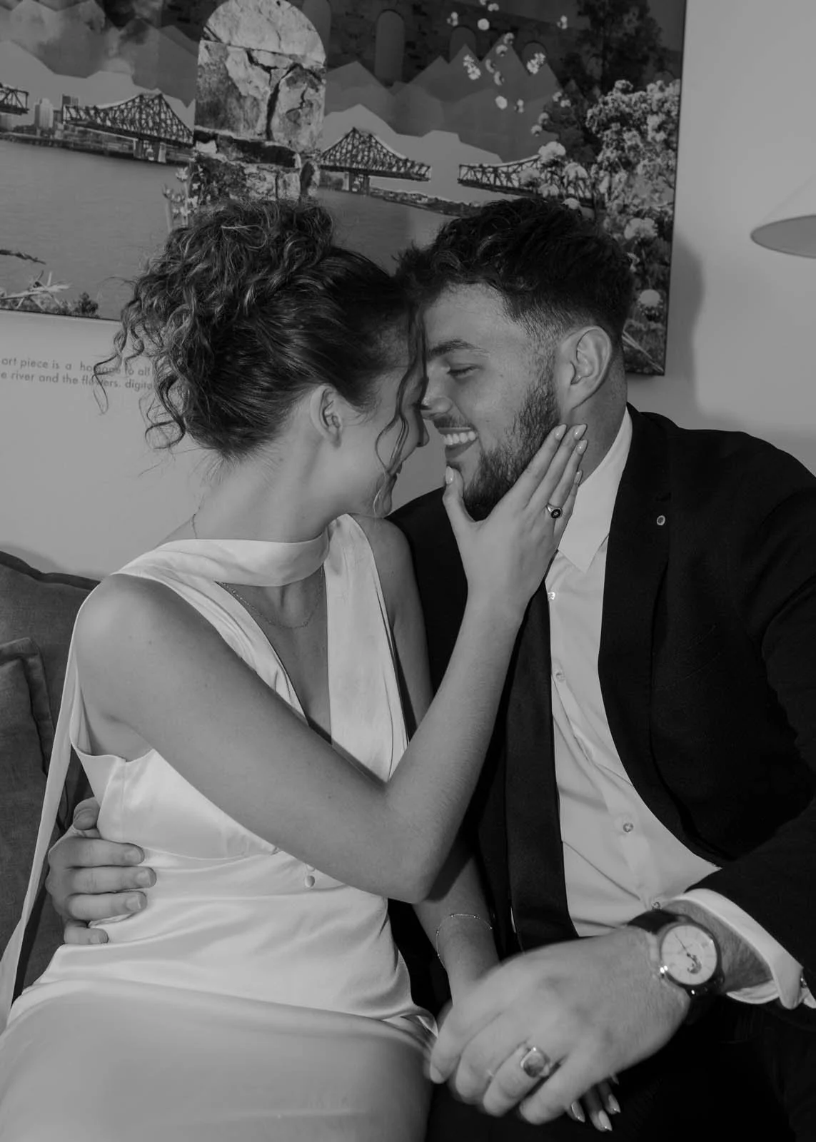 A black and white photo of a couple in a romantic moment, with faces close together, touching foreheads and smiling. The woman is wearing a sleeveless satin dress and has curly hair tied up, while the man is dressed in a suit with a watch and a ring on his finger.