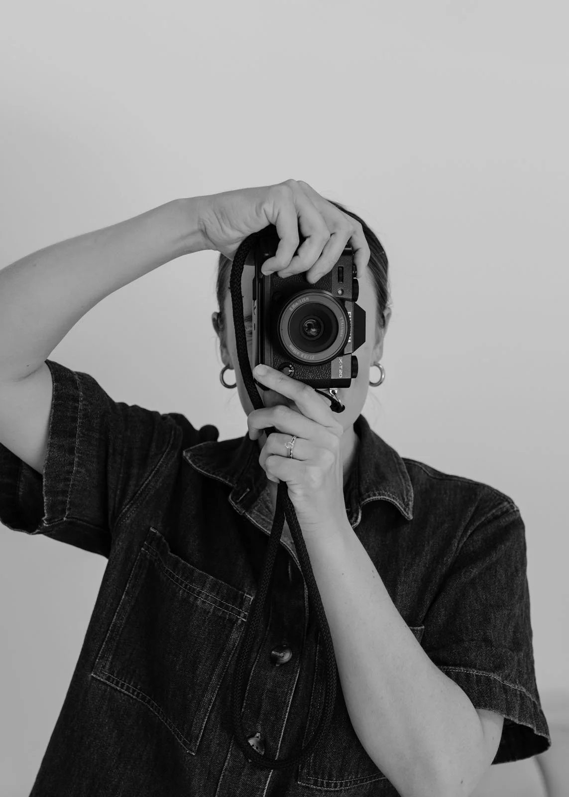 A person taking a selfie with a camera, holding it up to their face, in front of a plain background.