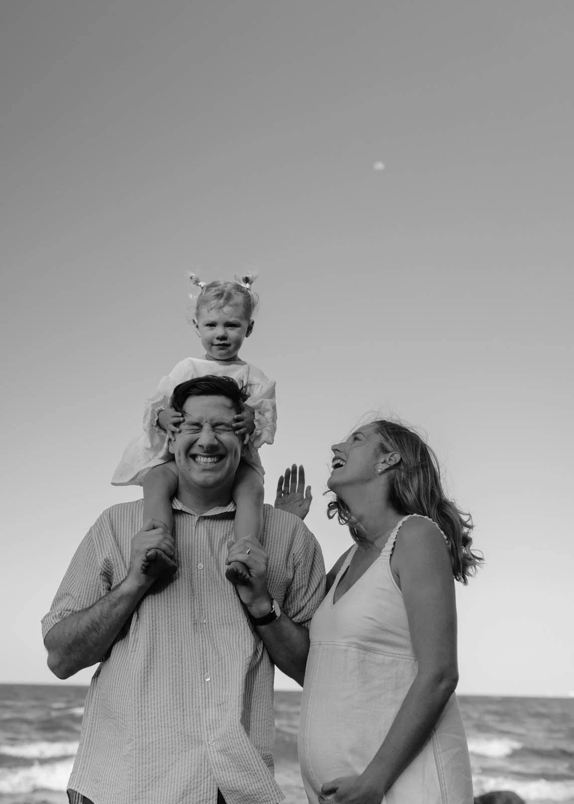 A black and white photo of a family at the beach: a man carrying a girl on his shoulders, a woman smiling and looking at them, with the ocean in the background and the moon in the sky.