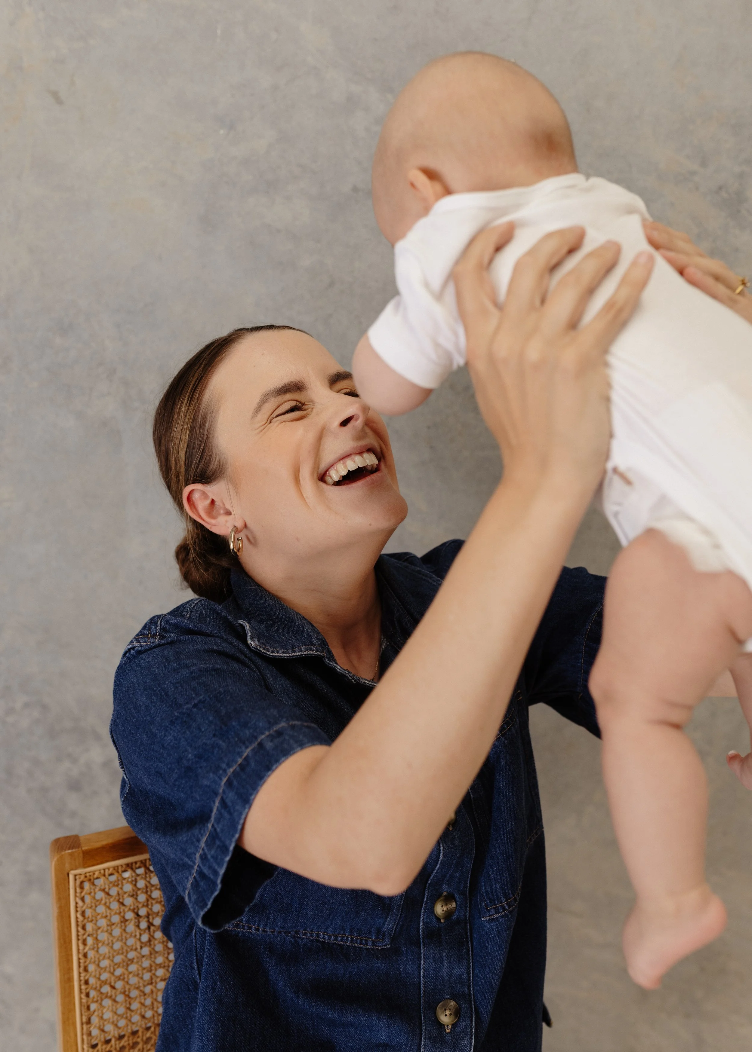 A woman with brown hair and earrings smiling happily while holding a smiling baby in a white outfit, against a neutral background.