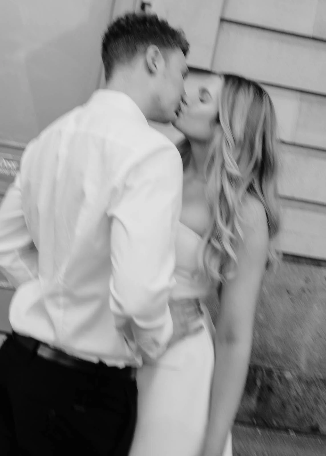 A black and white photo of a man and woman kissing, with the man wearing a white shirt and the woman wearing a strapless top, against a background of horizontal siding.