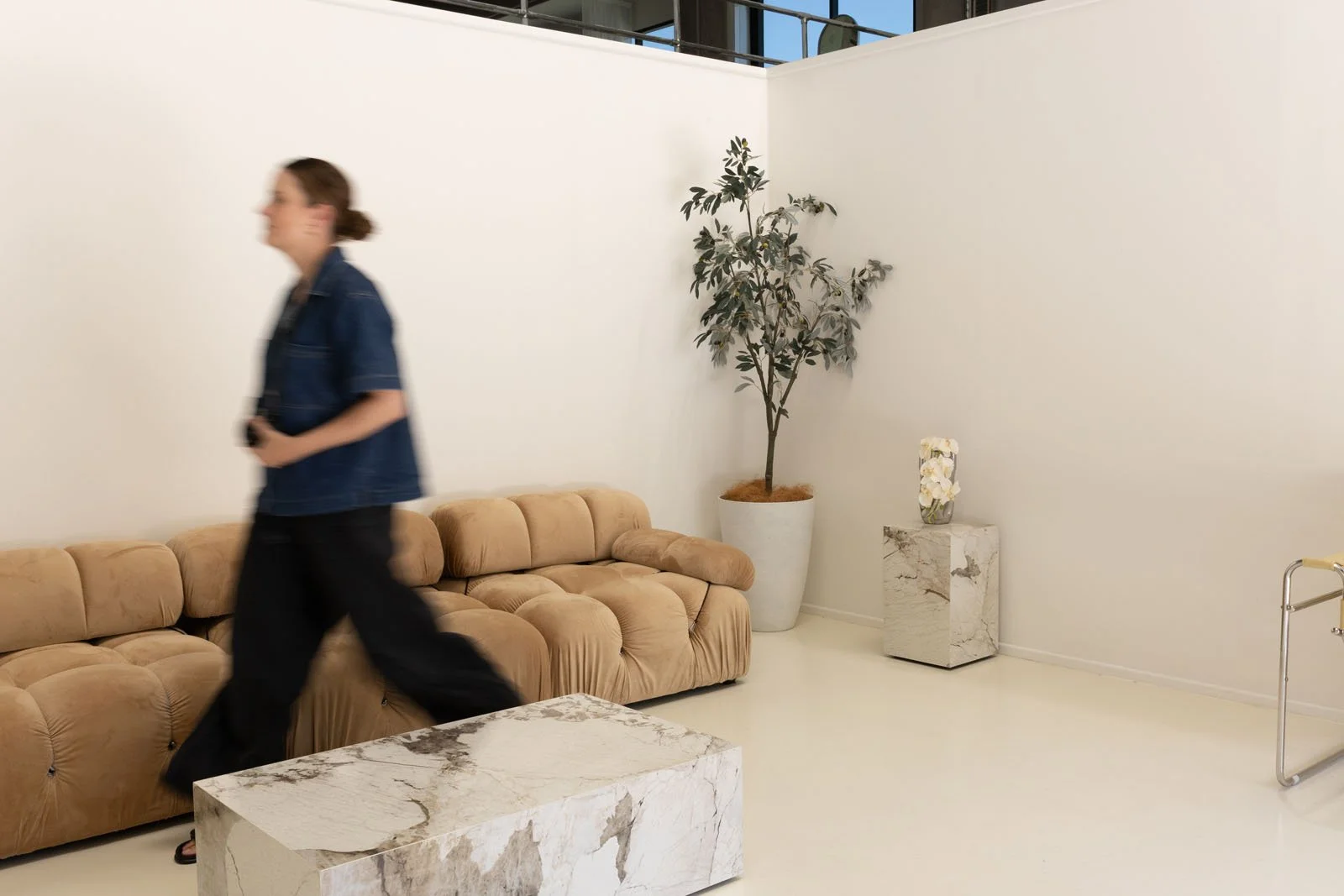 A woman walking in a minimalist living room with a beige couch, a small marble coffee table, a large potted tree, and a marble pedestal with a flower arrangement.