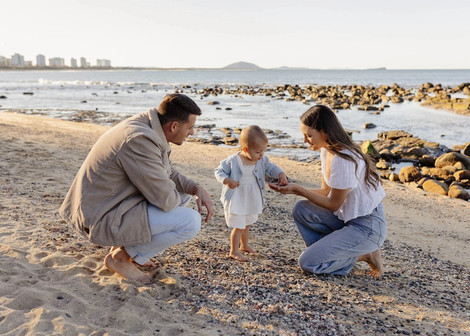A family of three, a man, a woman, and a young girl, on a beach looking at a shell together during sunset.