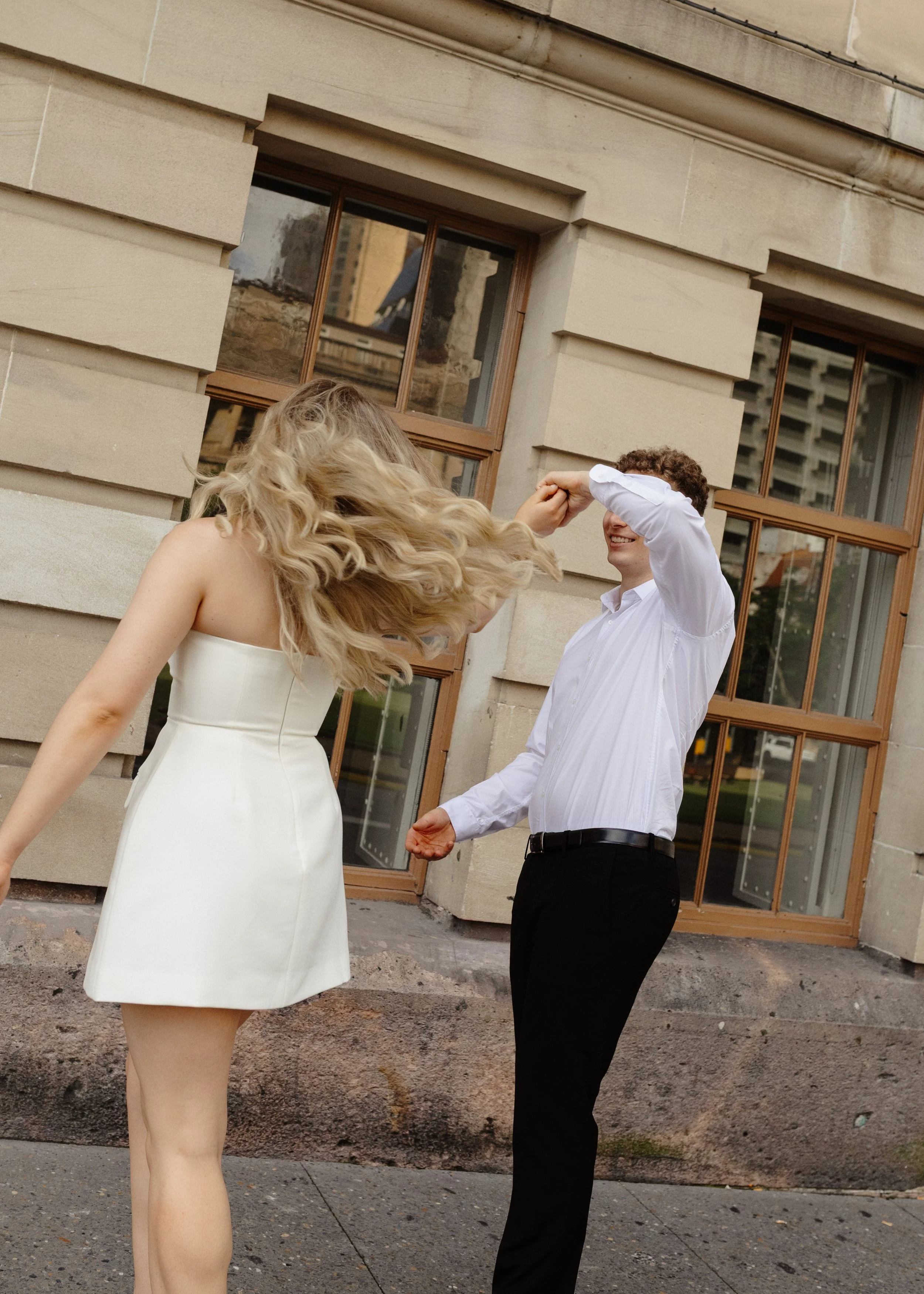 A man and woman dancing outdoors in front of a stone building with large windows, woman in a white dress, man in a white shirt and black pants.