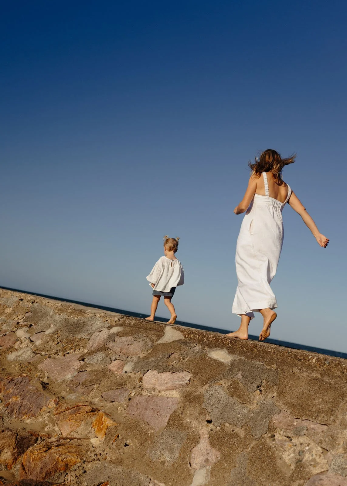 A woman and a young girl walking barefoot on a rocky surface outdoors, with a clear blue sky in the background.