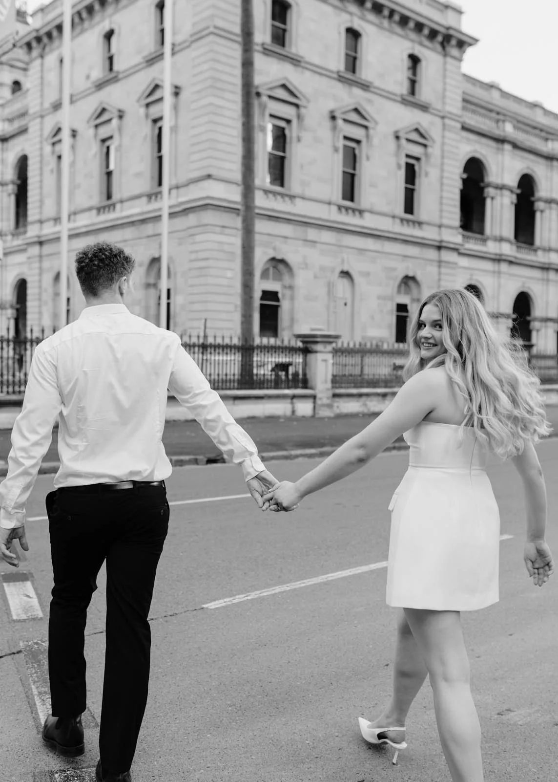 A black and white photo of a smiling woman and a man holding hands as they walk across the street in front of a historic building.