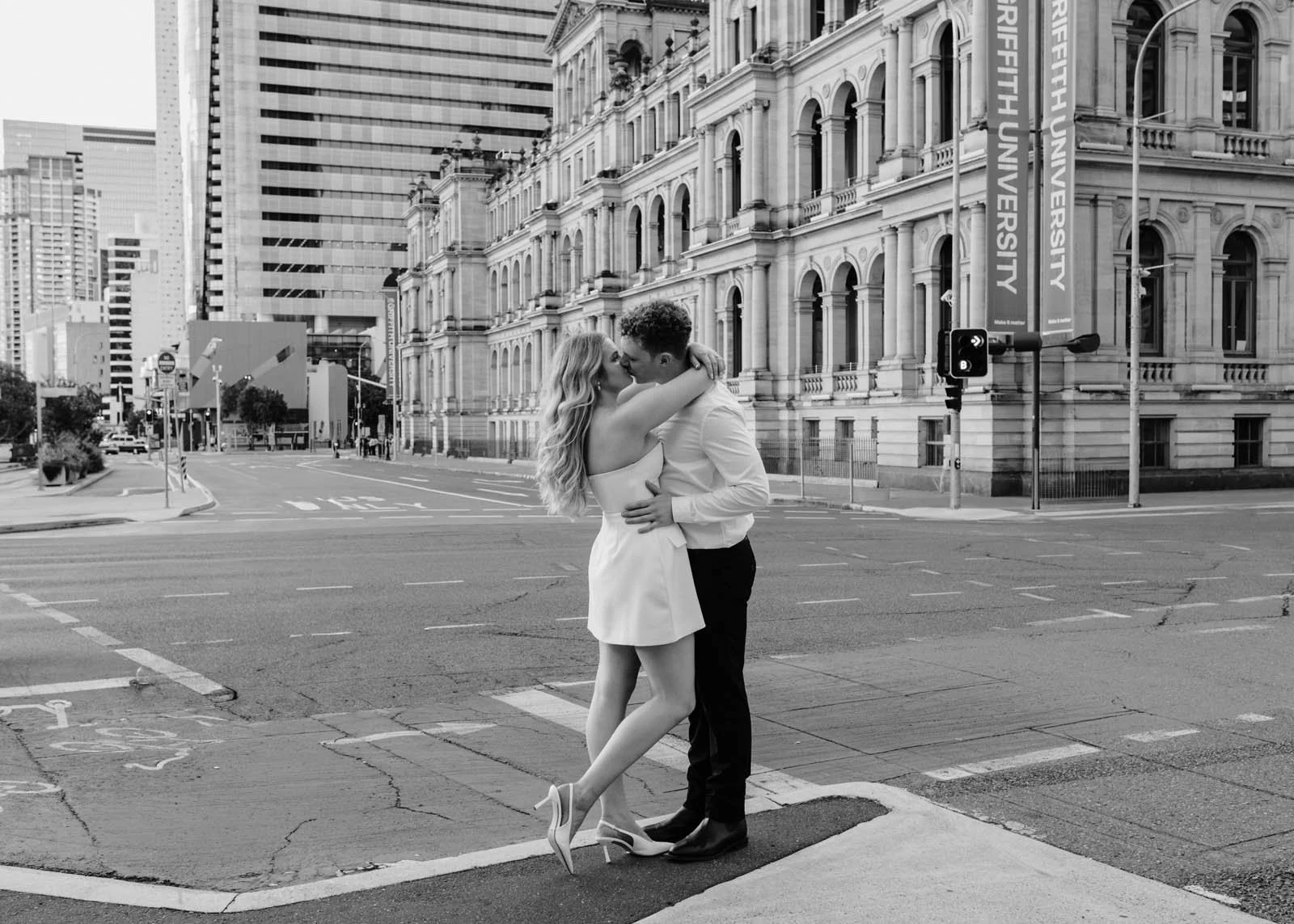 A black and white photo of a couple kissing on a city street corner, with buildings and traffic lights in the background. The woman is wearing a short dress and high heels, and the man is dressed in a shirt and pants.