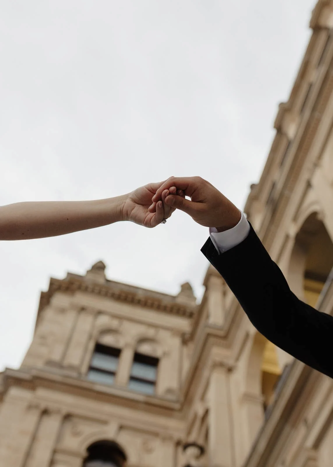 A close-up of a man in a tuxedo holding hands with a woman, against a backdrop of a historic building and an overcast sky.