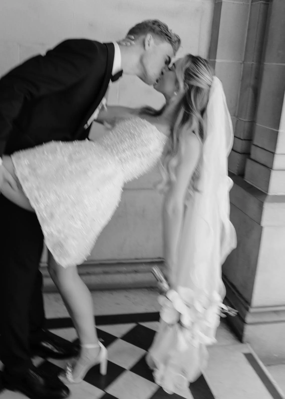 A newlywed couple kissing at their wedding reception, the groom in a black tuxedo and the bride in a striking white wedding dress with high heels, in a black and white photo.