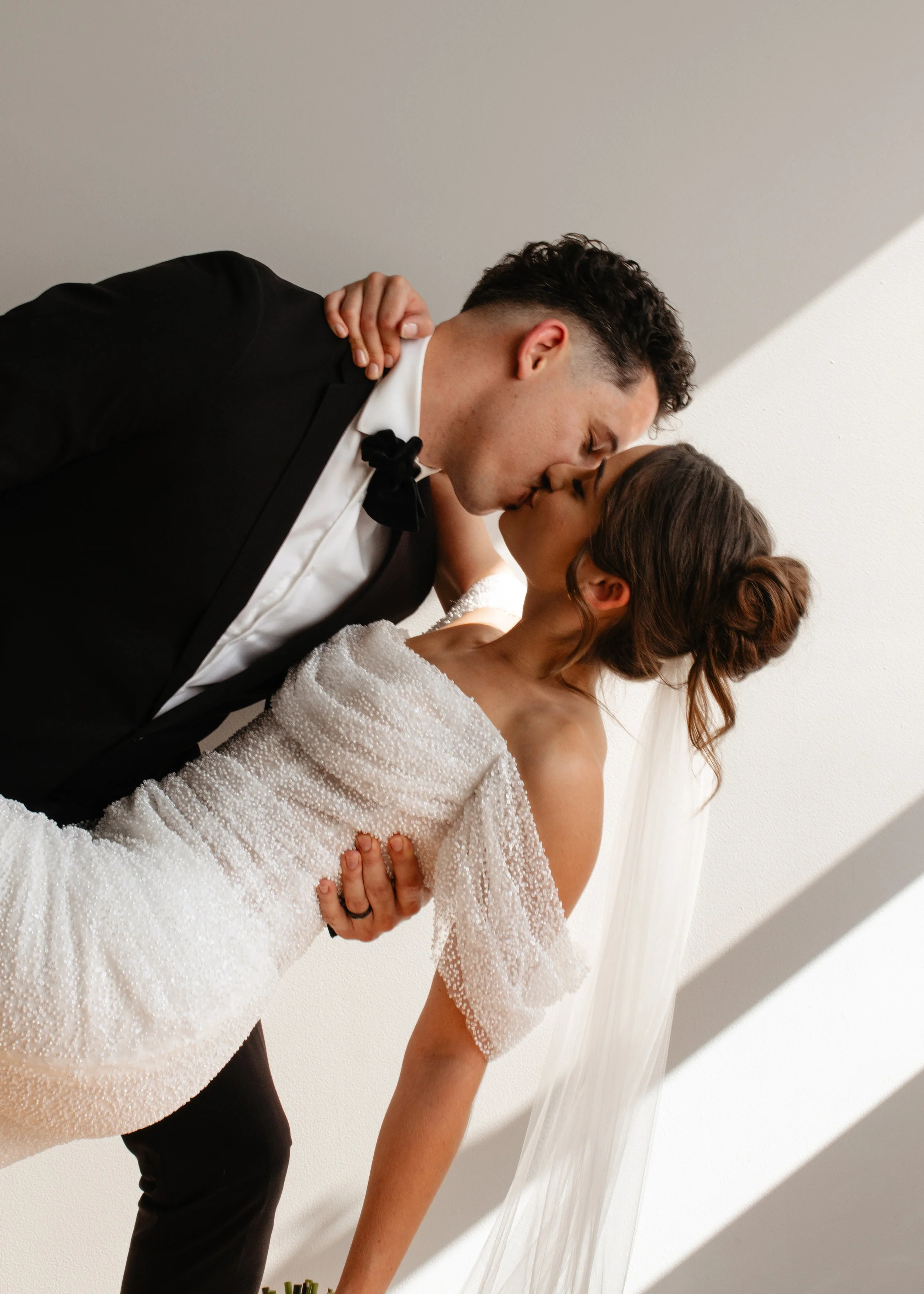A bride and groom sharing a kiss during their wedding, with the groom in a tuxedo and the bride in a white beaded gown and veil.