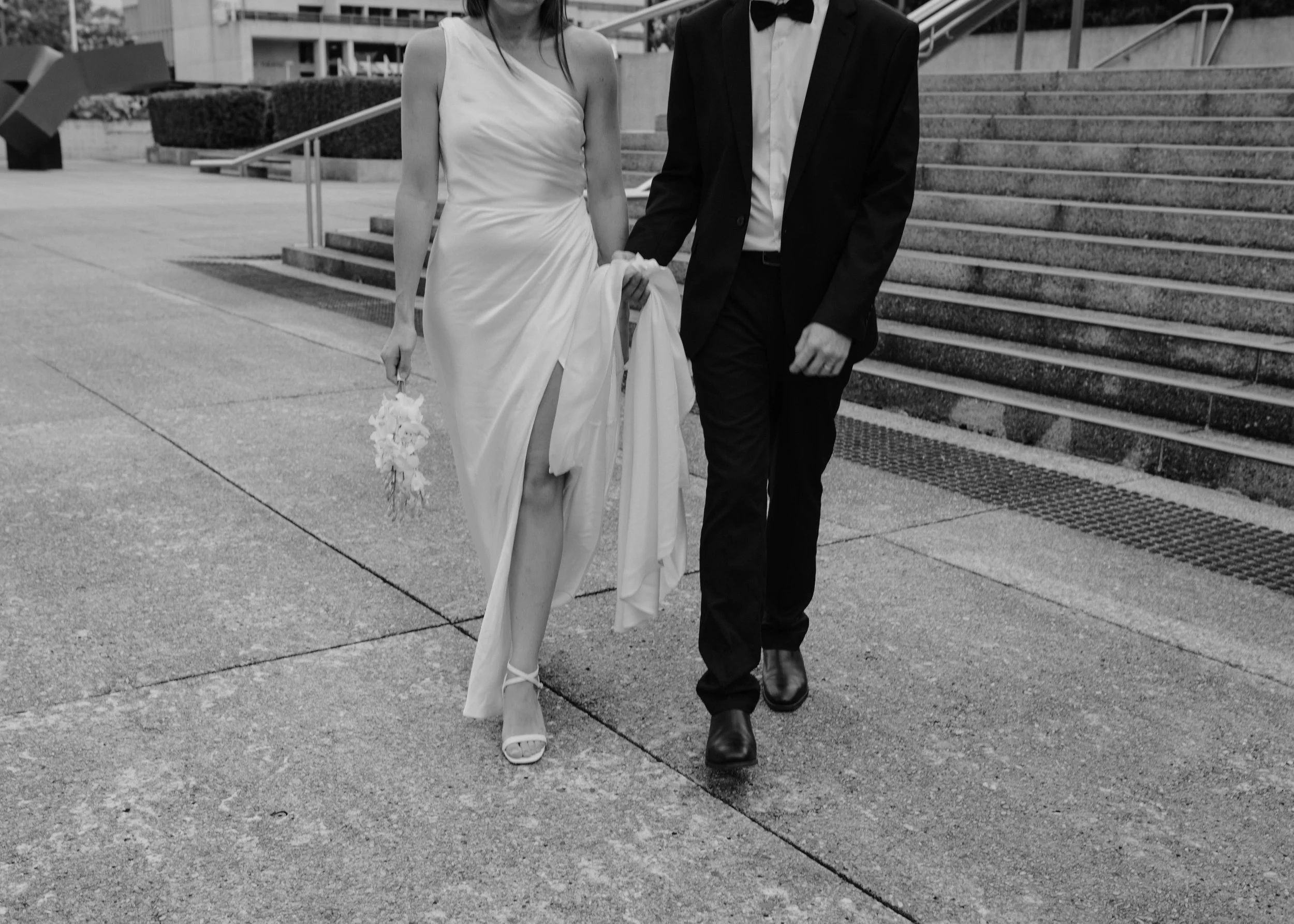 A black and white photo of a couple in wedding attire walking outdoors. The bride is wearing a satin wedding gown with a thigh-high slit, strappy heels, and holding a small bouquet. The groom is dressed in a tuxedo with a bow tie, holding the bride's