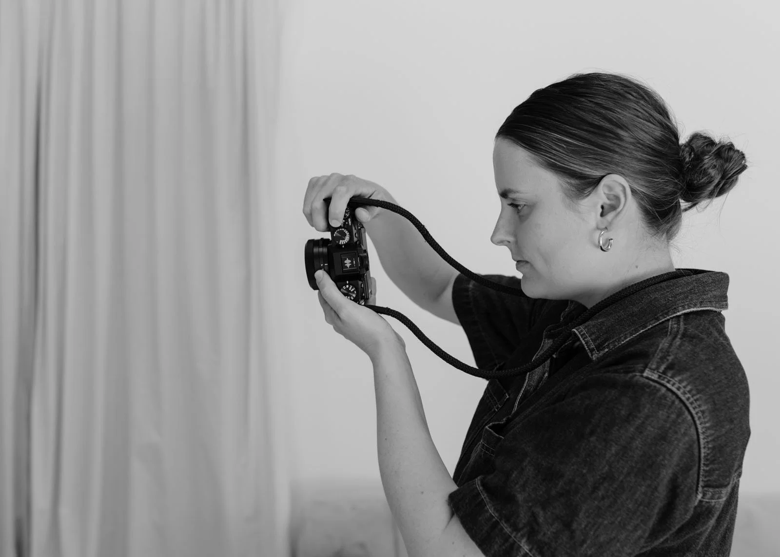 A woman takes a photo with a digital camera, with her hair tied back and wearing a denim jacket, indoors against a plain wall with curtains.
