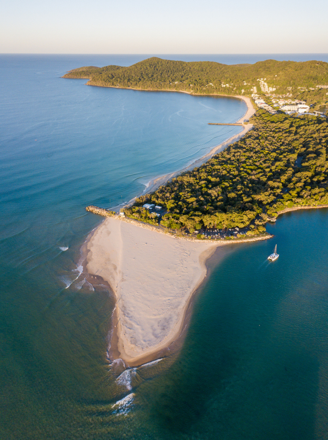 An aerial view of a coastal area featuring a sandy beach, a small boat on the water, green forested hills, and buildings in the background.