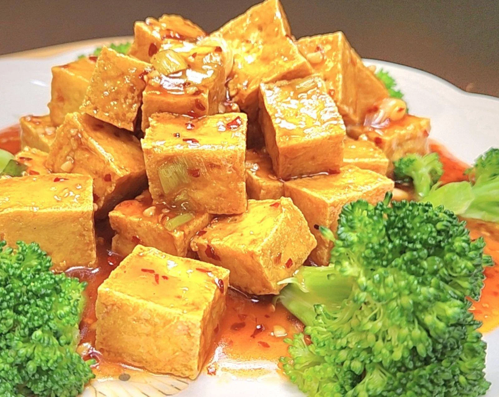 A dish with cubed, fried tofu topped with a savory sauce, garnished with chopped green onions, and served with broccoli and other vegetables.