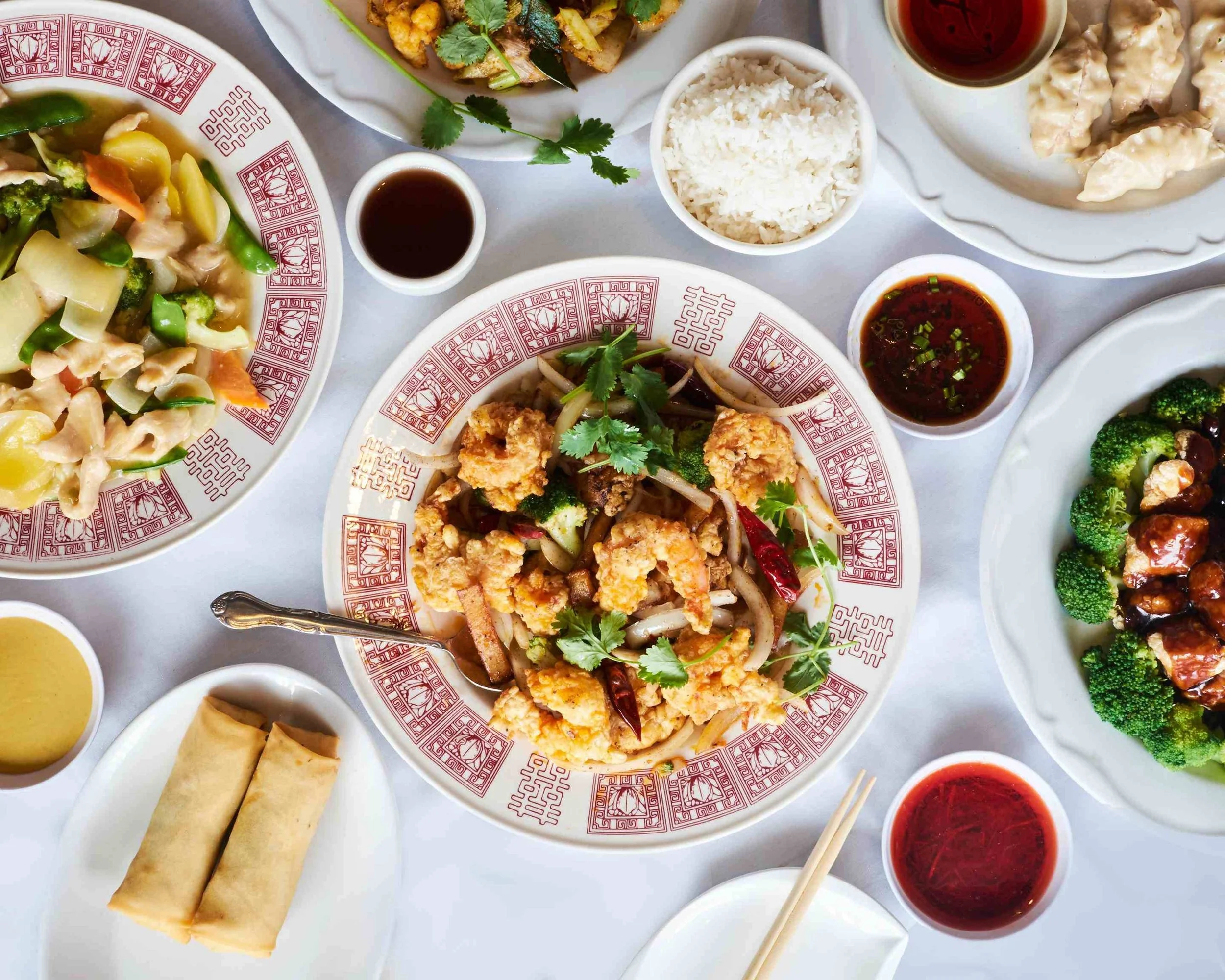 Assorted Chinese dishes on a white table, including stir-fried vegetables, fried chicken with broccoli, spring rolls, white rice, dumplings, and various dipping sauces.