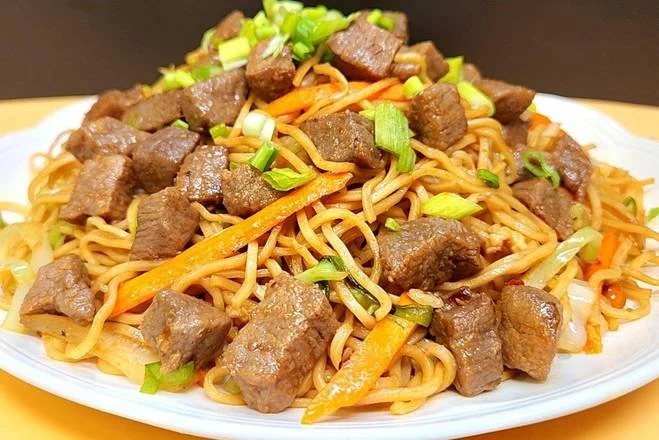 Plate of stir-fried noodles topped with cooked beef cubes, sliced green onions, and shredded carrots.