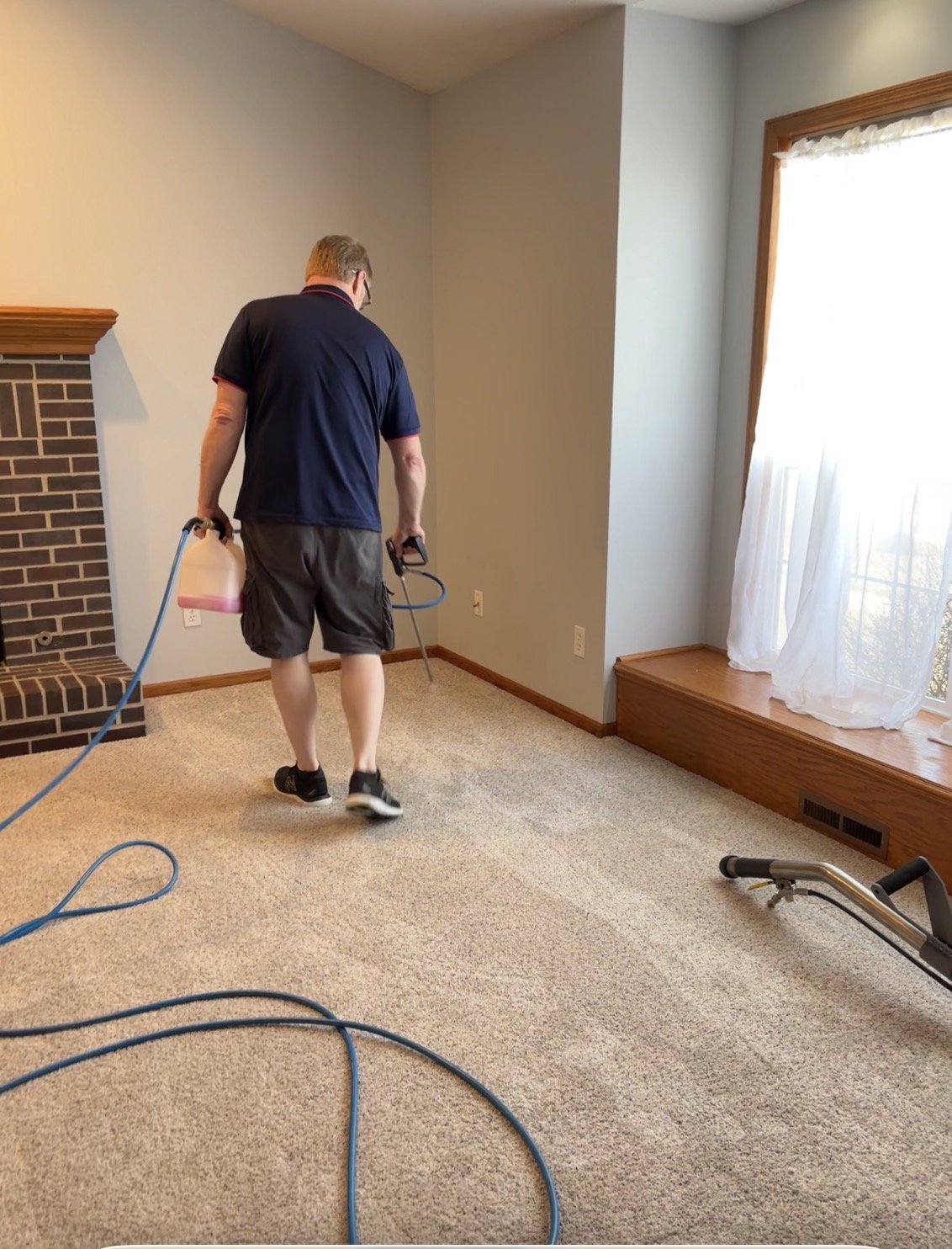 A man is cleaning beige carpet in a living room with a handheld carpet cleaning machine. The room has a brick fireplace, a large window with white curtains, and a row of wooden trim at the base of the wall.