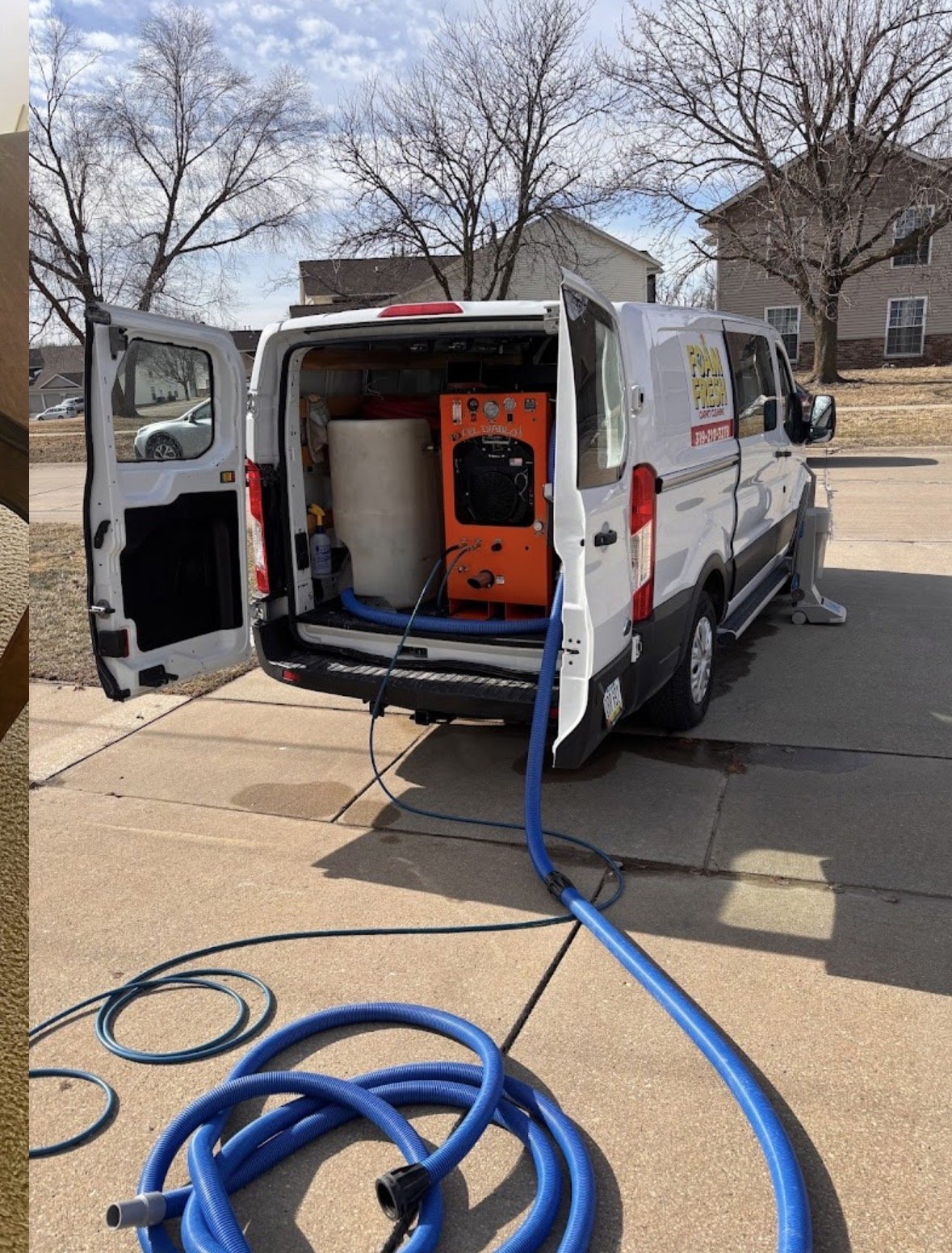 A van with open rear doors parked on a driveway, containing equipment for cleaning or washing, with blue hoses extending from it. The background shows residential houses and leafless trees on a partly cloudy day.