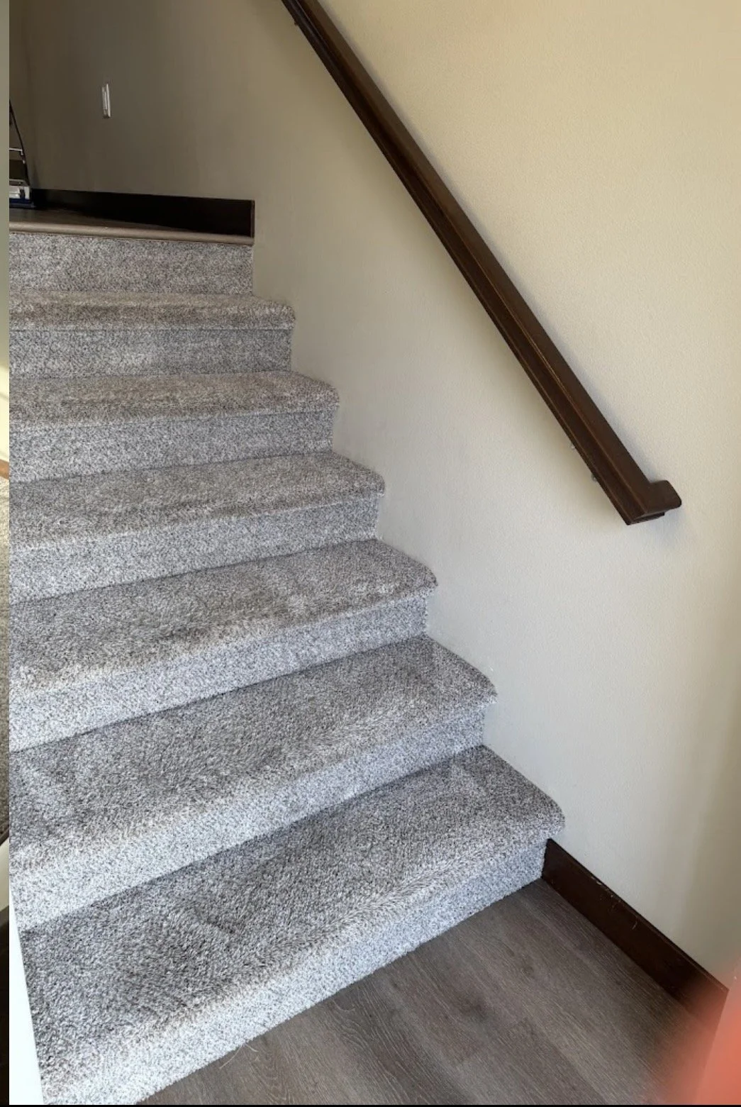 Carpeted staircase with a wooden handrail on the right side, next to a beige wall and hardwood flooring at the bottom.