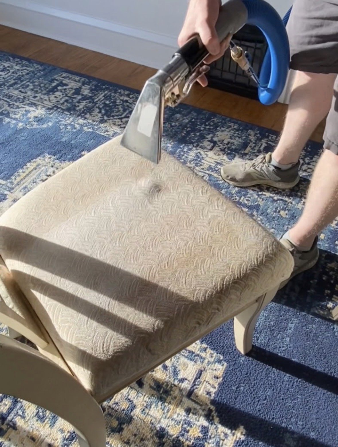 Person cleaning a beige upholstered chair with a handheld steam cleaner indoors, with a blue and cream patterned rug visible.
