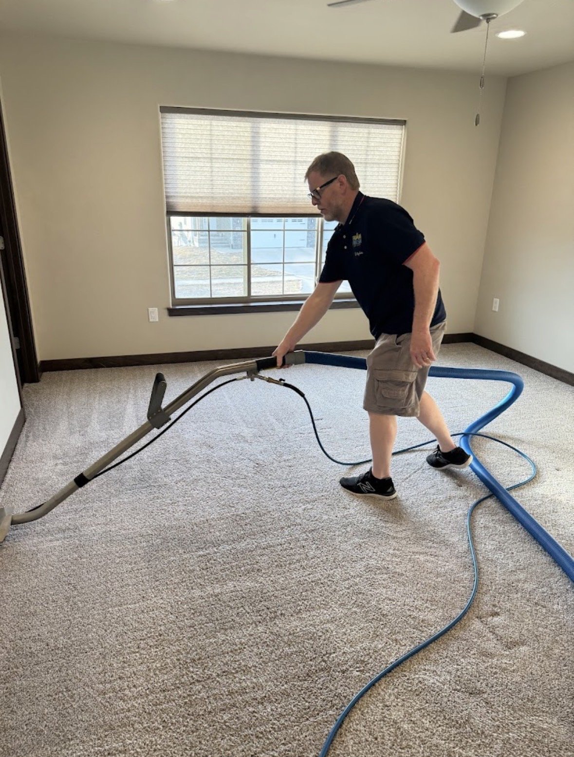 A man cleaning a beige carpet in a room with a carpet cleaning machine, a large window, and beige walls.