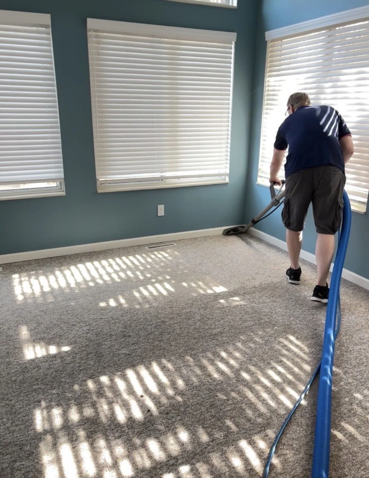 Person vacuuming light-colored carpet in a room with blue walls and three large windows with white blinds.