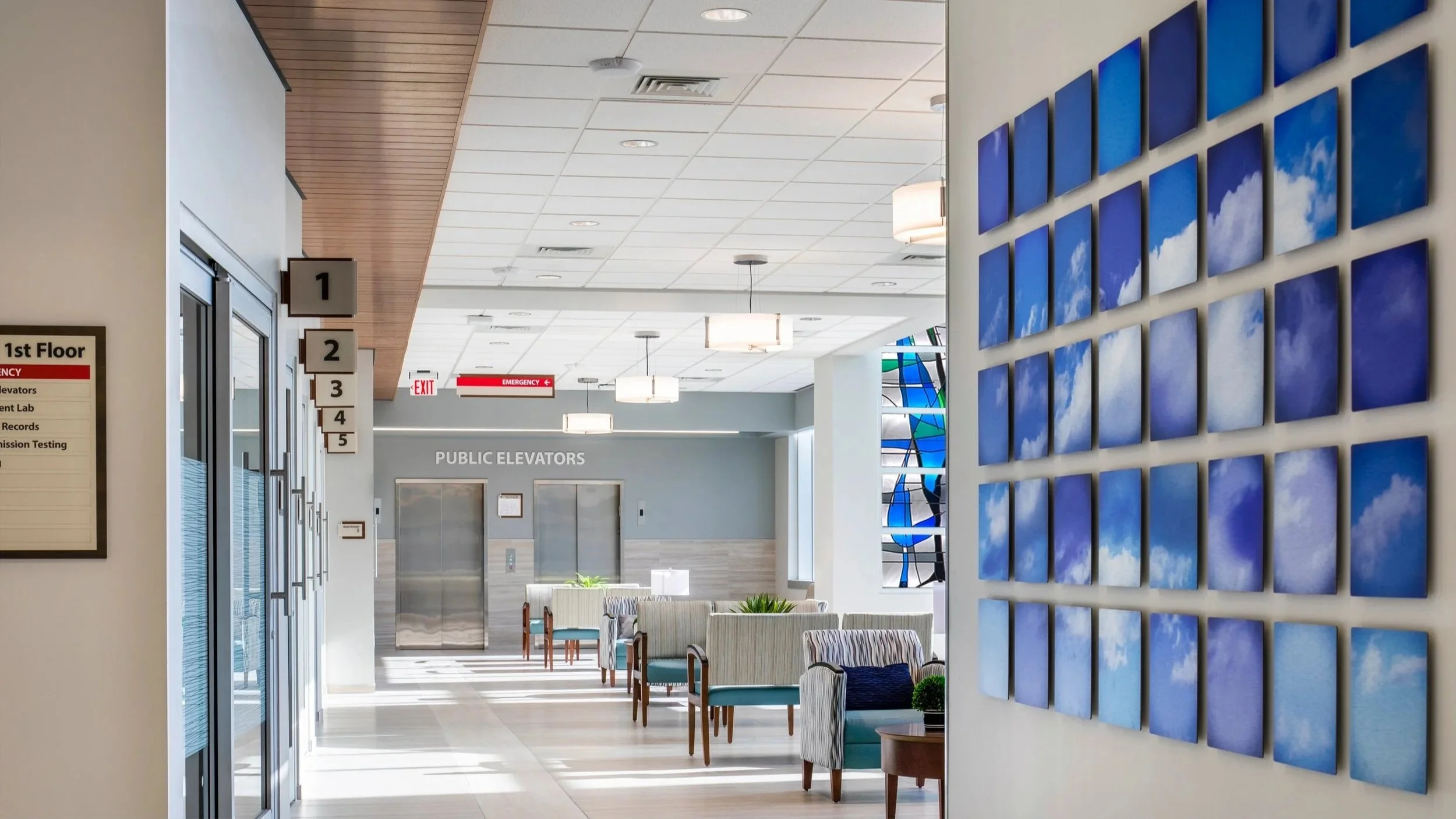 Interior of a modern building lobby with seating area and elevator doors, featuring wall art of blue sky and clouds, with signs indicating floors and emergency exit.