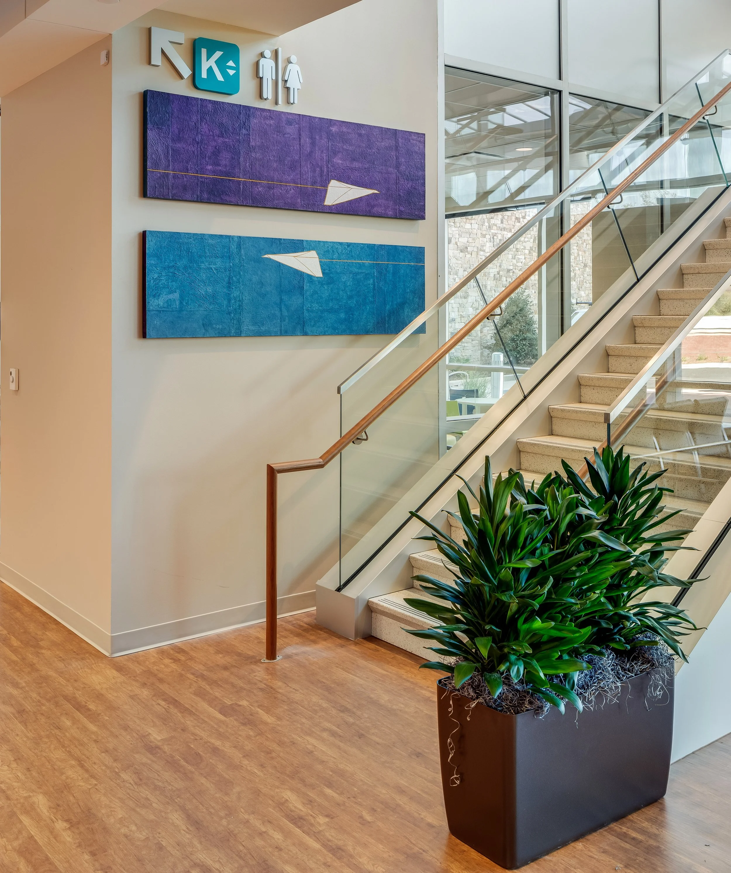 Indoor area with wooden flooring, beige wall, large potted green plant, staircase with glass railing, artwork on wall, bathroom sign with male and female symbols, and directional arrow
