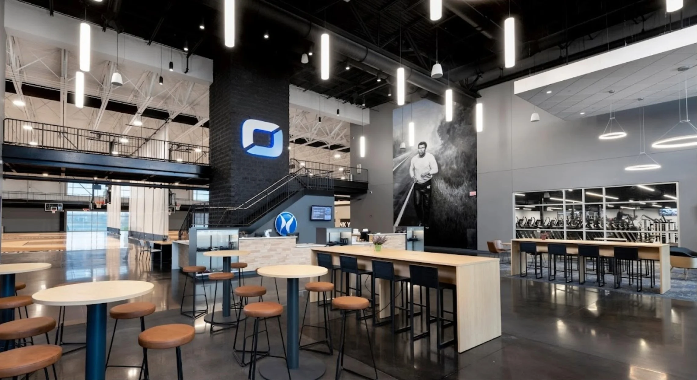 Interior of a modern sports or fitness center with a high ceiling, bright lighting, and multiple basketball hoops. There's a reception area with tables and chairs, a large black-and-white portrait of a man running on a trail, and the center of the sp