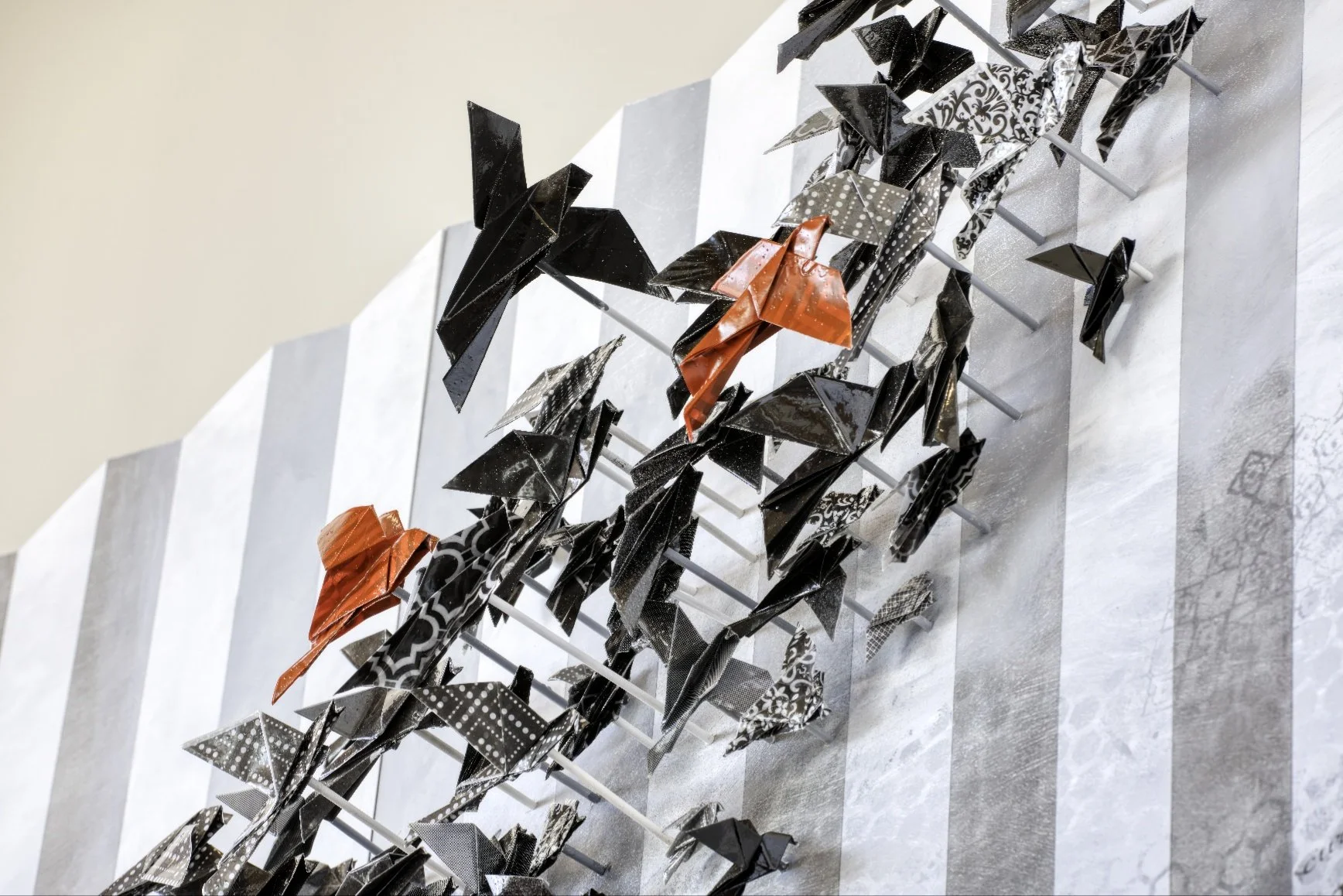 An art installation featuring black and red origami paper birds mounted on white sticks, displayed on a marble surface with a marble-patterned wall in the background.