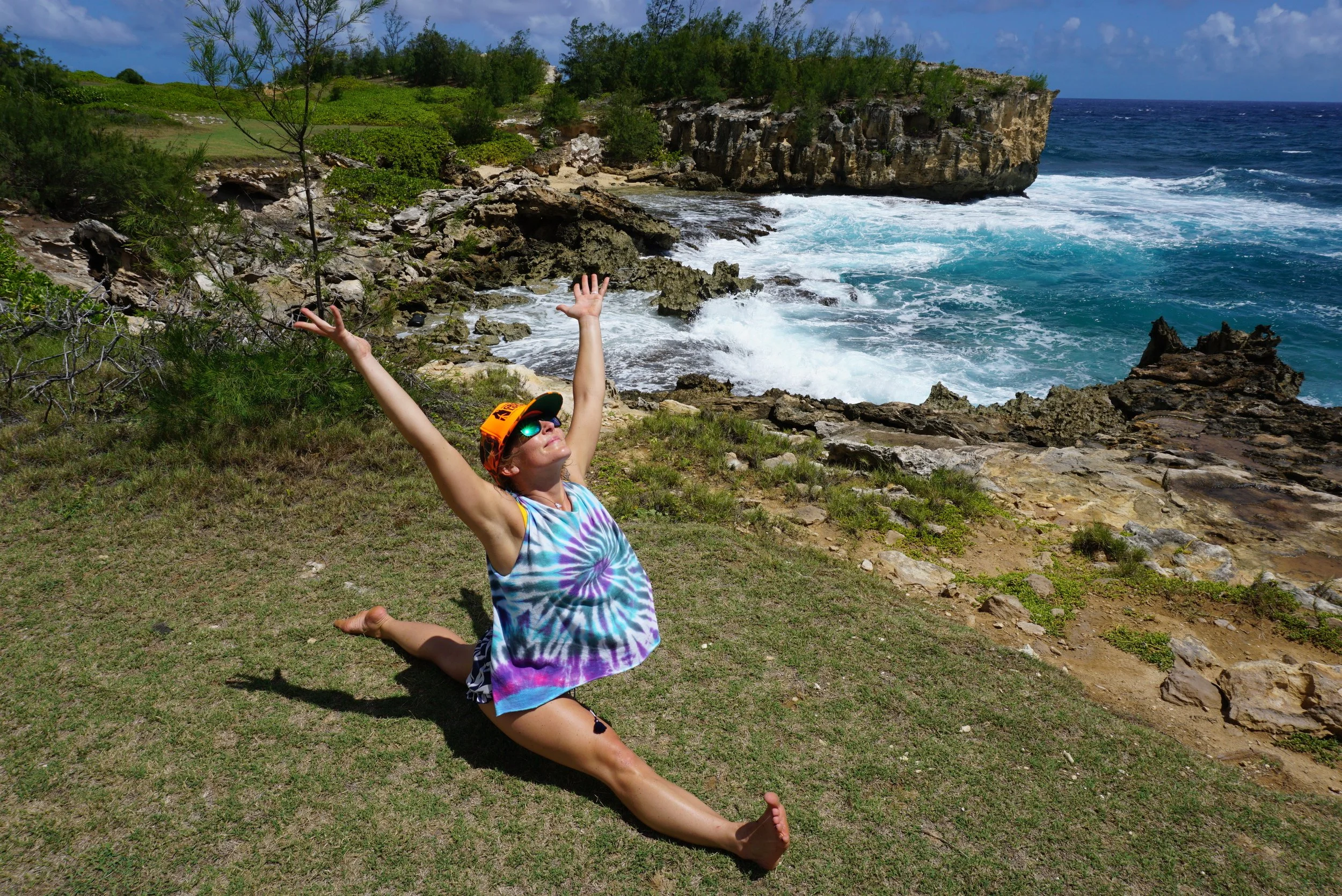 A woman performing a split yoga pose on grass near the rocky shoreline of the ocean, wearing a tie-dye tank top, a colorful cap, sunglasses, and a bracelet, with waves crashing on rocks in the background.
