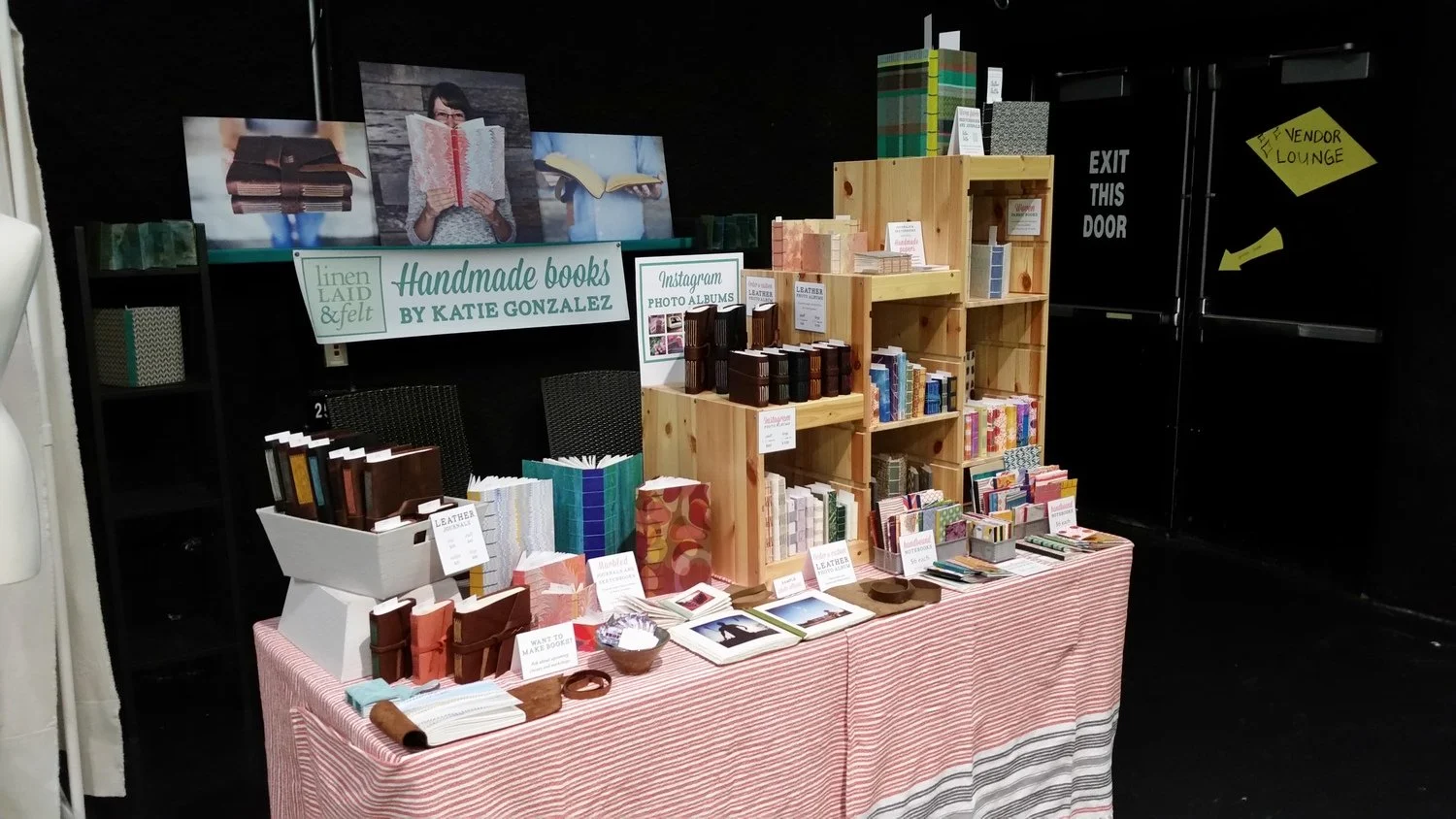 Display of handmade books and leather goods at a craft fair stall, with various books, leather wallets, and photo albums arranged on a table with a red and white striped tablecloth. Signage indicates handmade books by Katie Gonzalez and Instagram photo albums.
