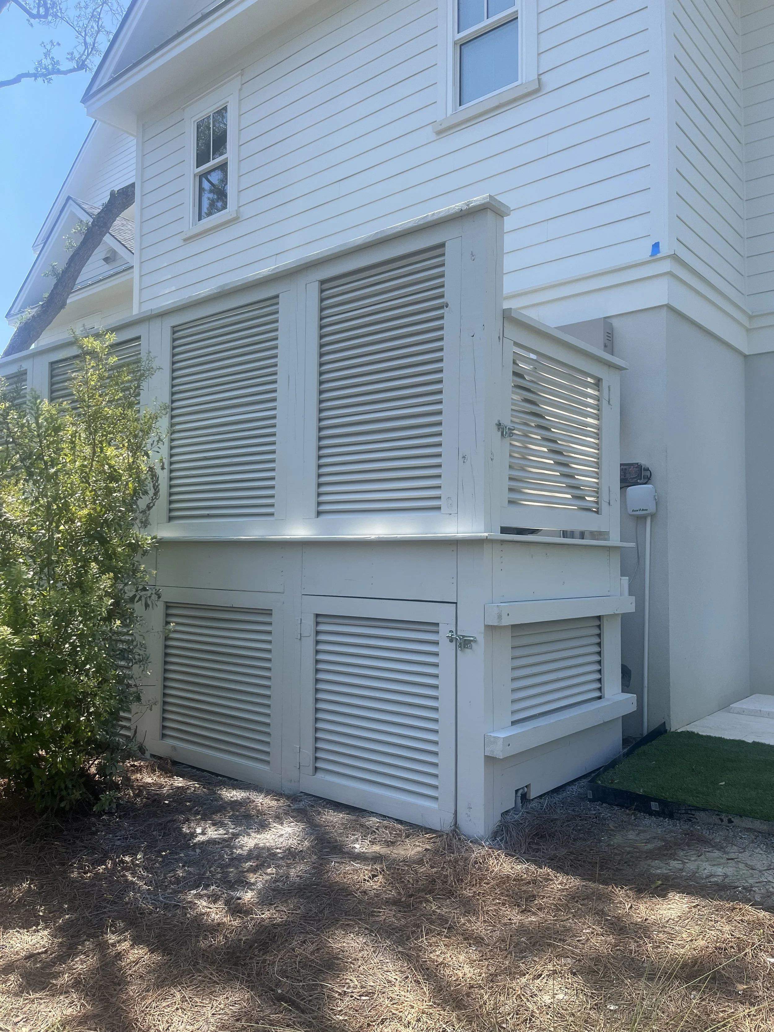 The image shows the side of a white house with siding, featuring two windows on the upper level and a vented enclosure on the lower level. There are some trees and bushes nearby, with the ground covered in dried grass and dirt.