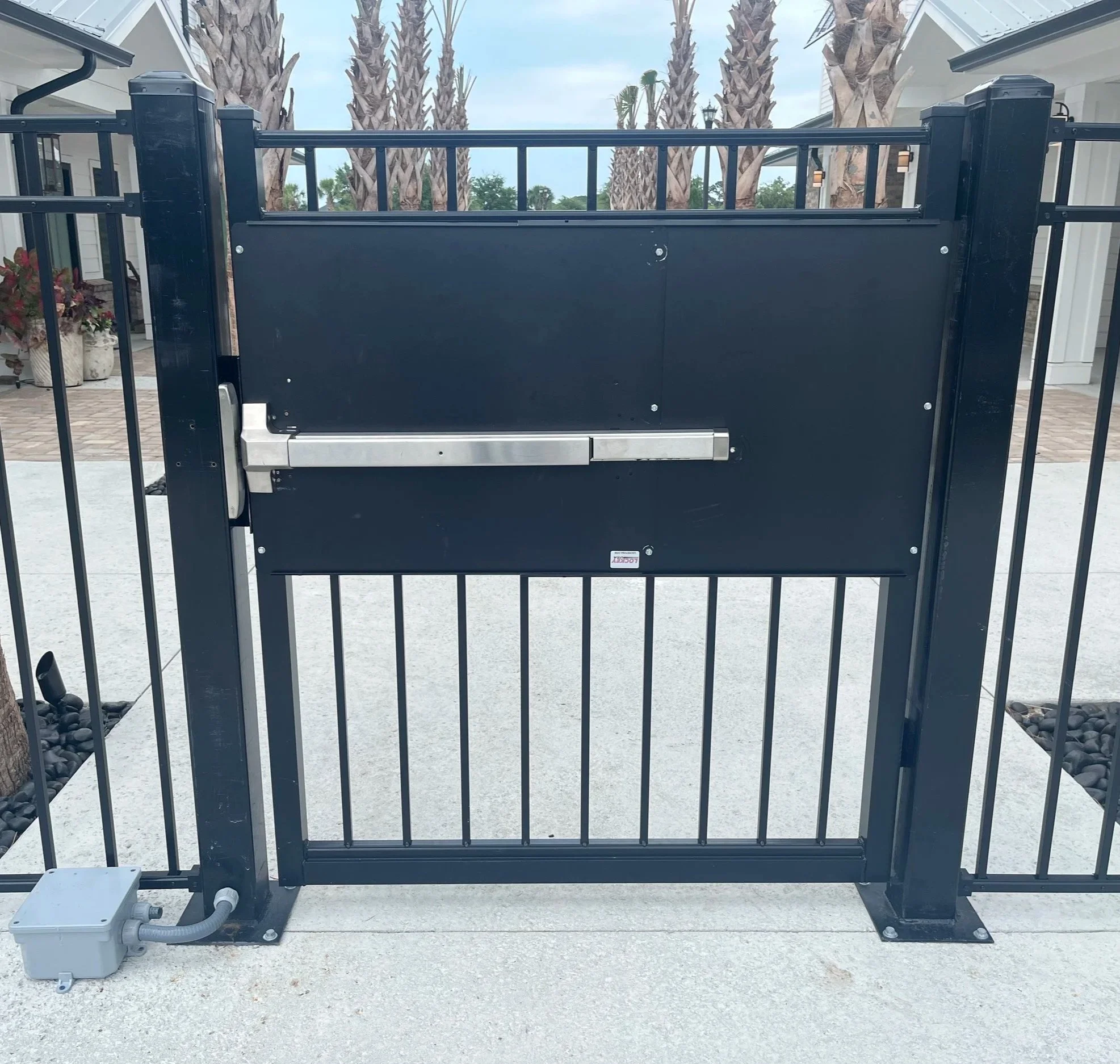 Electric gate with hardware component at the bottom left, black metal fence, palm trees and a partly cloudy sky in the background.