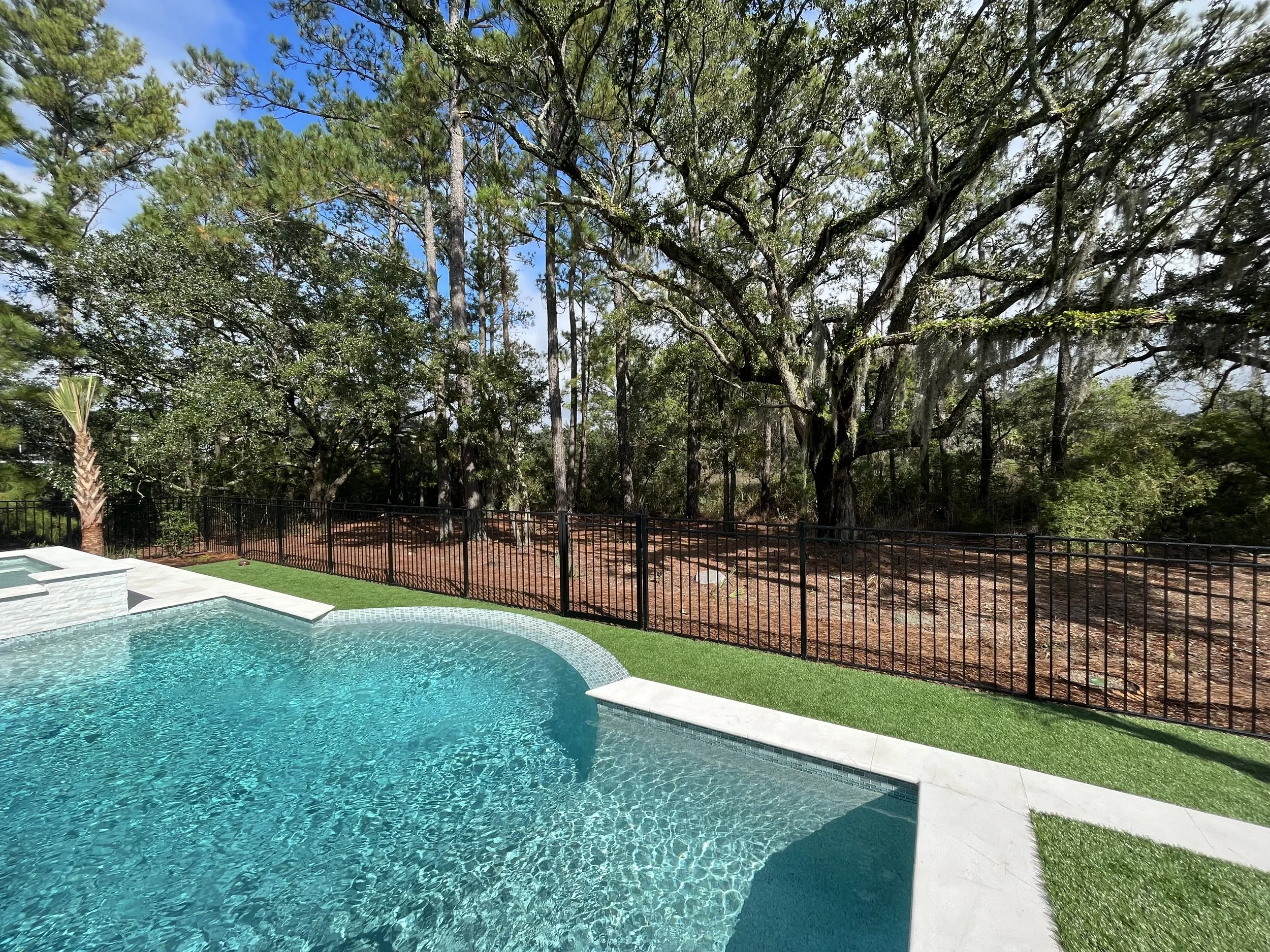 A backyard swimming pool with a curved edge, surrounded by a green artificial grass area and a black aluminum fence. Beyond the fence are tall trees with green leaves and blue sky with some clouds.