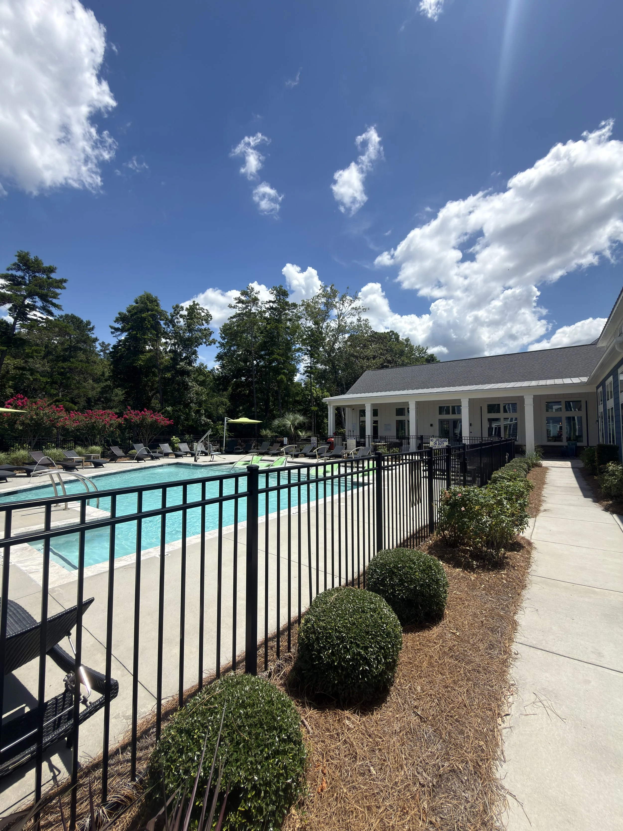 A backyard with a swimming pool surrounded by lounge chairs, a black aluminum fence, a building with a porch, green bushes, and trees under a partly cloudy sky.