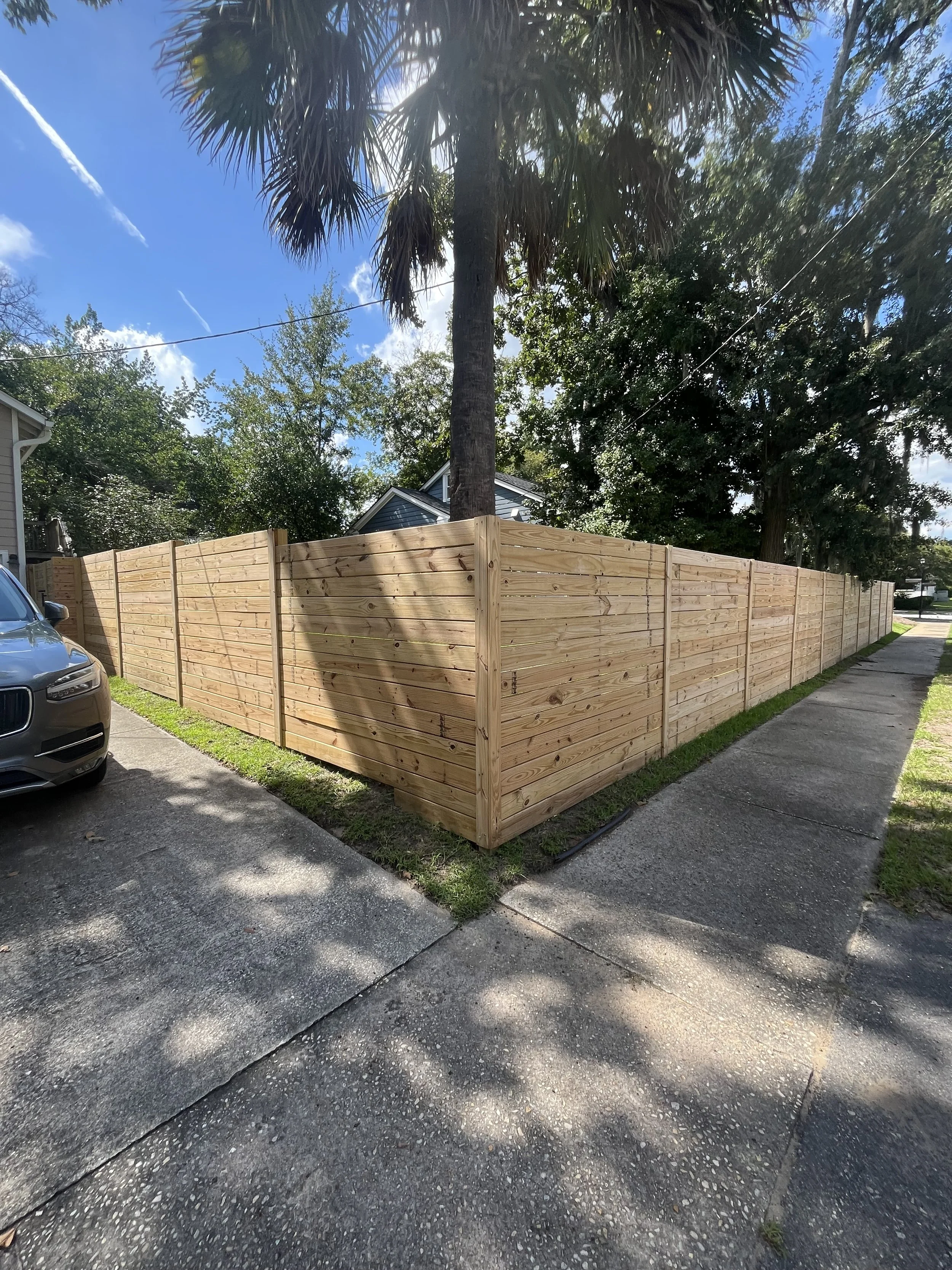 A newly constructed wooden fence surrounds a backyard, with a large palm tree and other trees providing shade. The fence runs along a sidewalk and a parked car is partially visible in the foreground. The sky is partly cloudy with sunlight filtering through the trees.