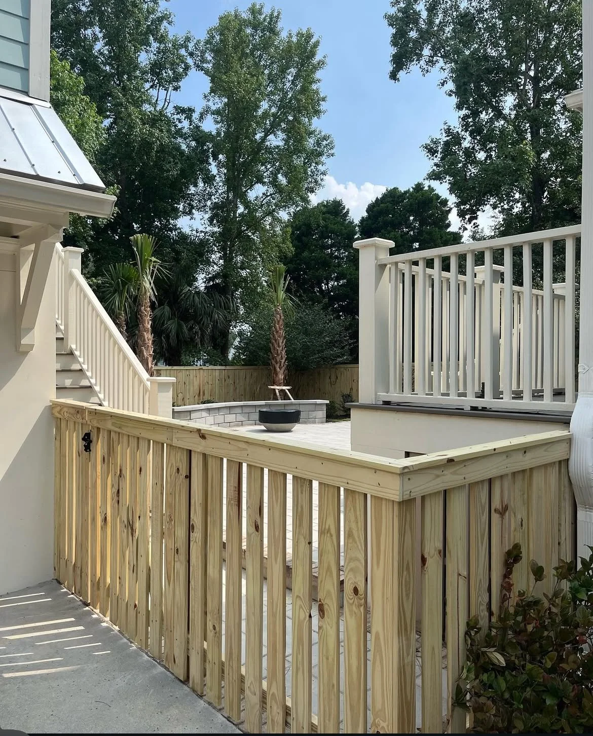 A backyard patio area with new wooden fencing, a staircase, and lush green trees in the background.
