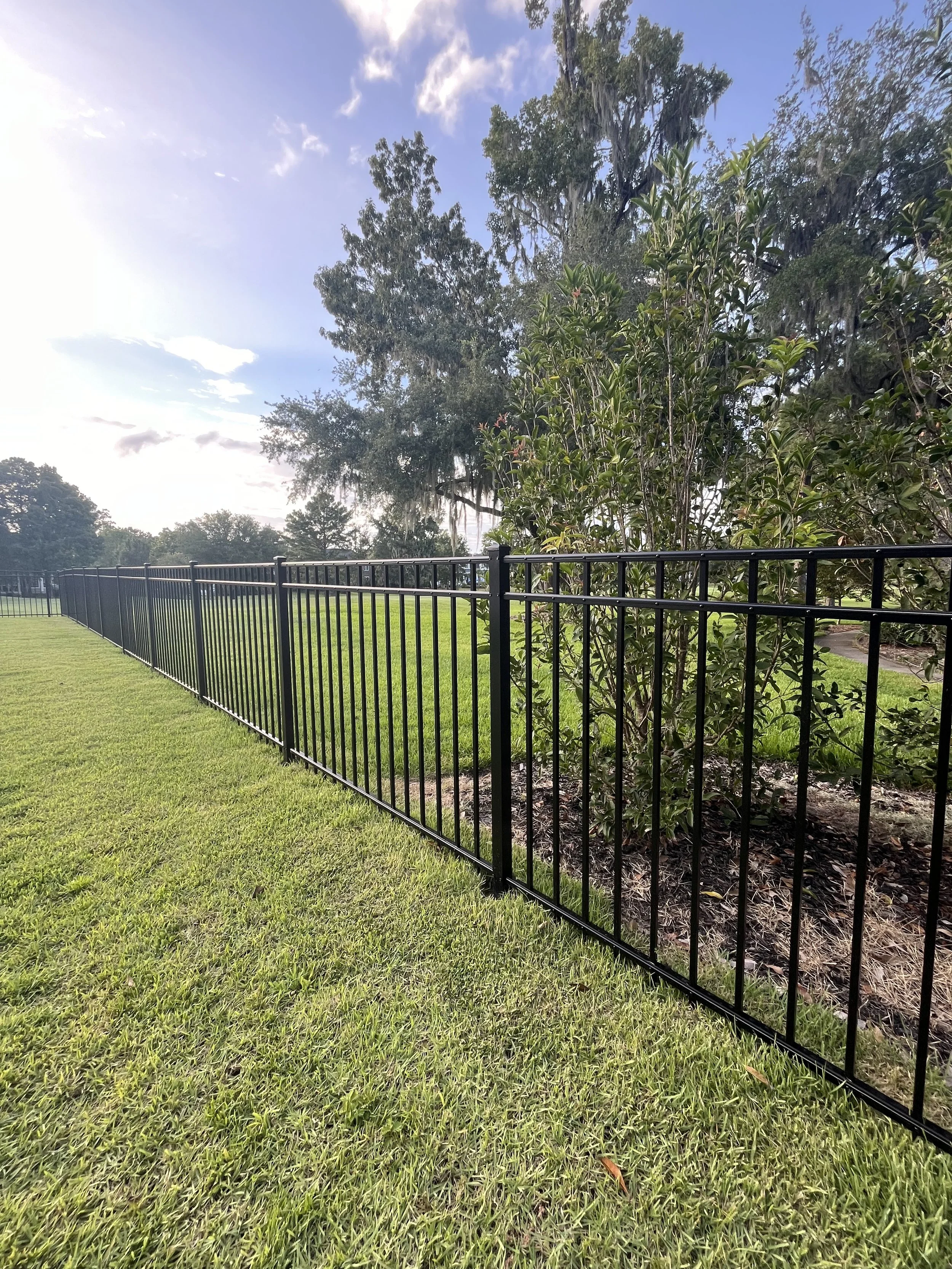 A black aluminum fence runs along a well-maintained grassy park area with tall trees in the background. The sky is partly cloudy with patches of blue visible.