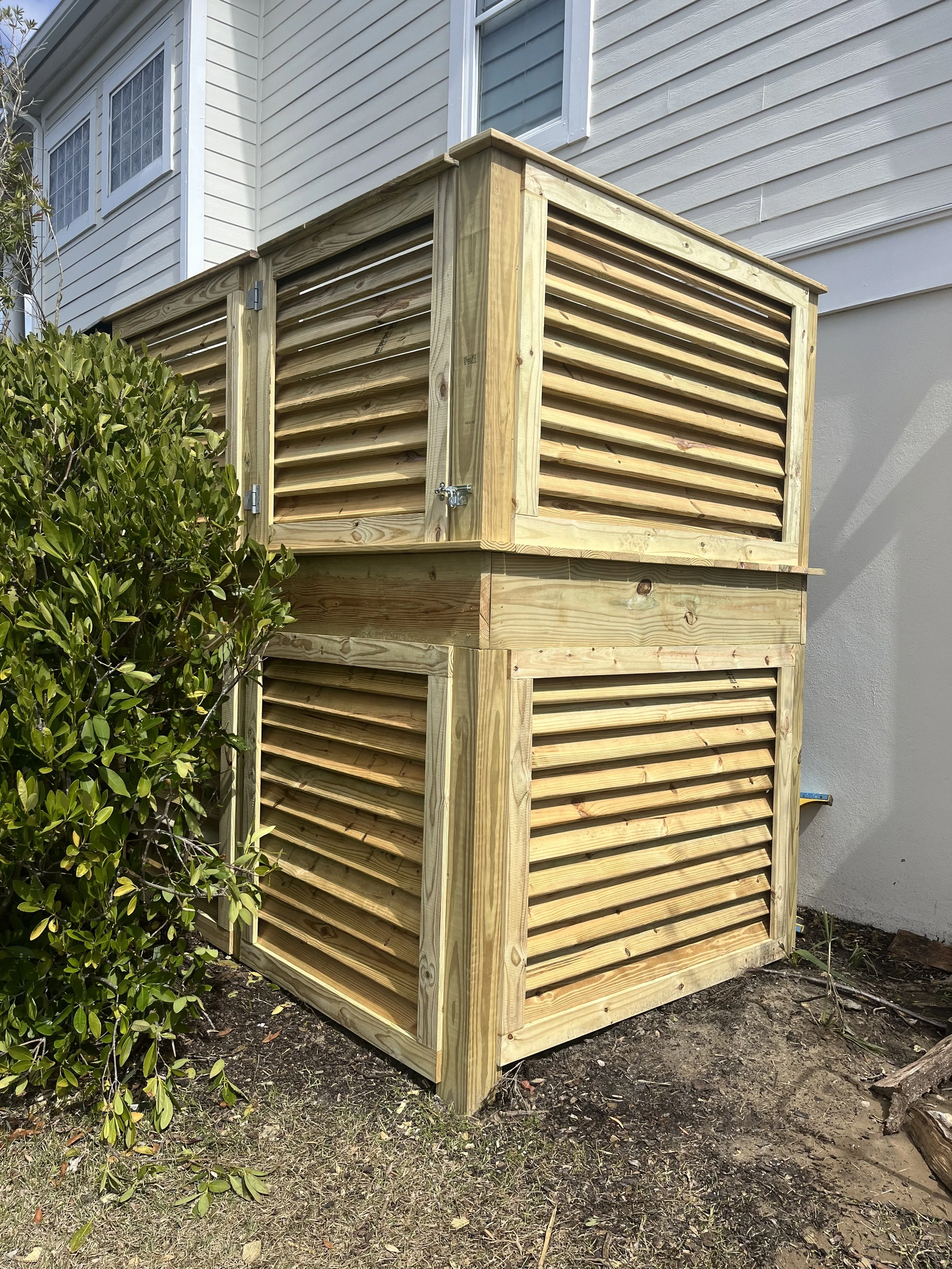 A two-story wooden structure with horizontal slatted sides, installed next to a white house with horizontal siding and a window. The ground below is dirt with some small plants and leaves.
