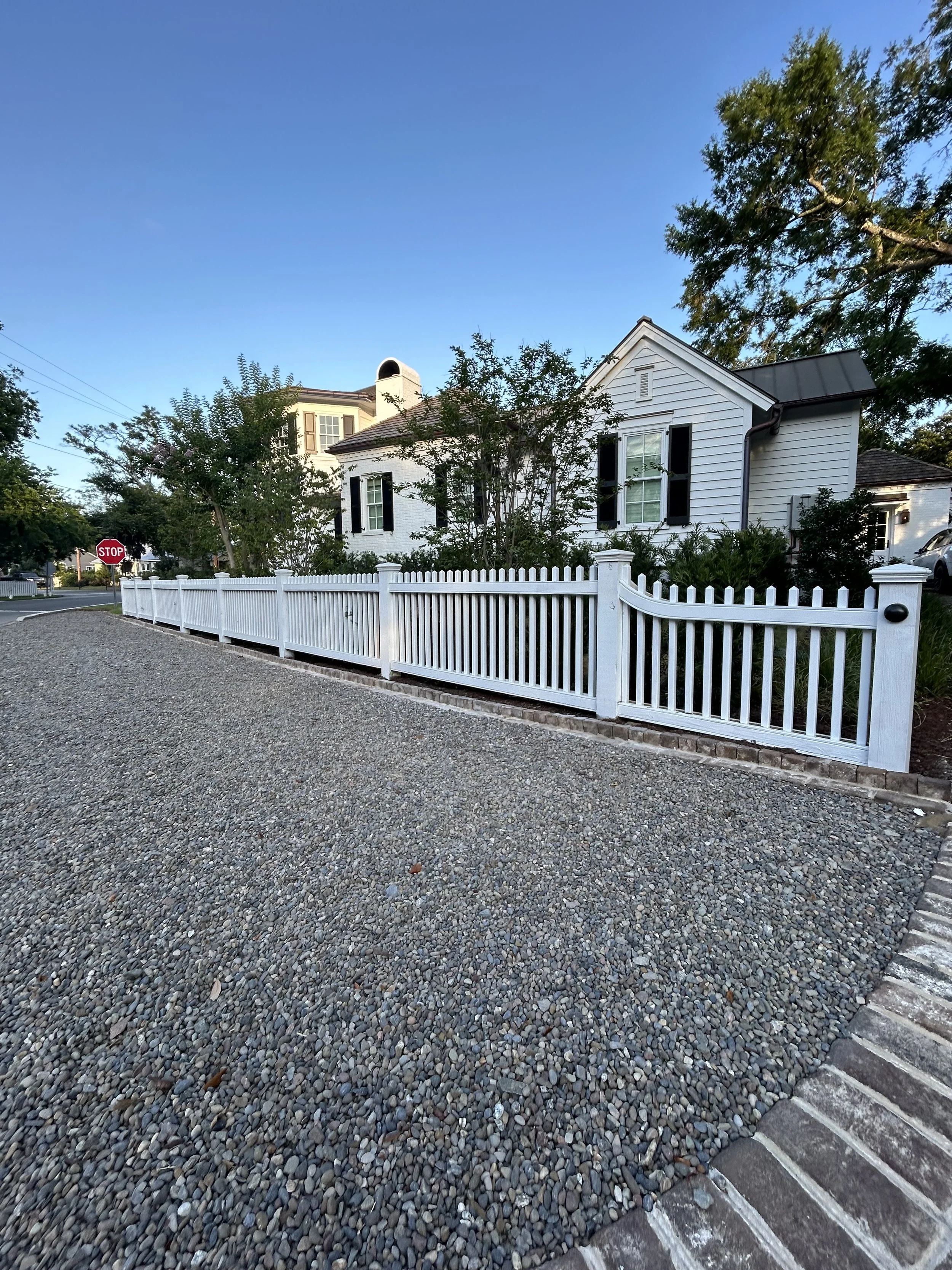 A white picket fence surrounds a front yard with trees and bushes in front of a white house with black shutters. A gravel driveway is in the foreground, and a stop sign is at the street corner. The sky is clear and blue.