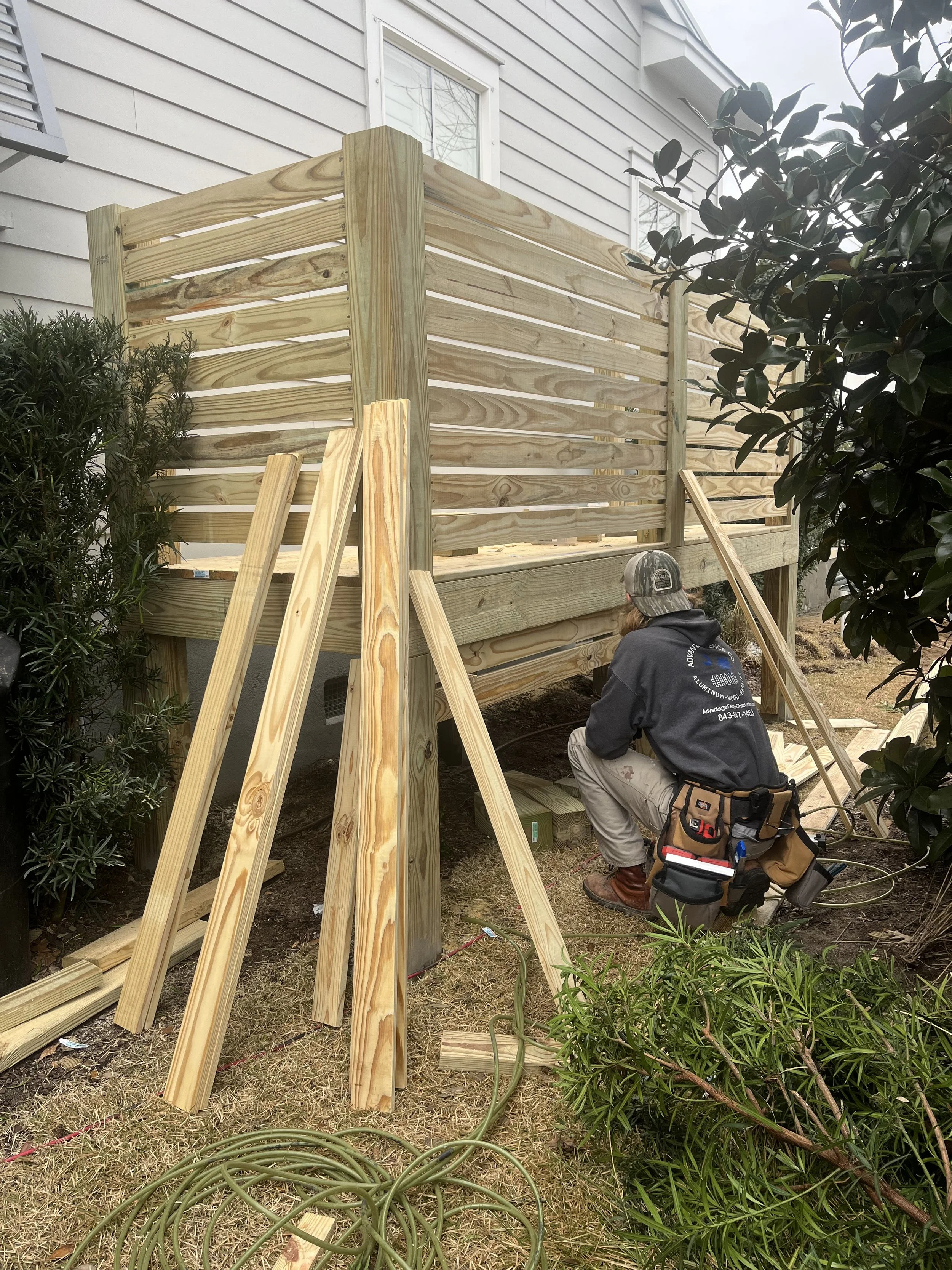 A person working on building a wooden deck or fence outside a house, with tools and lumber around.